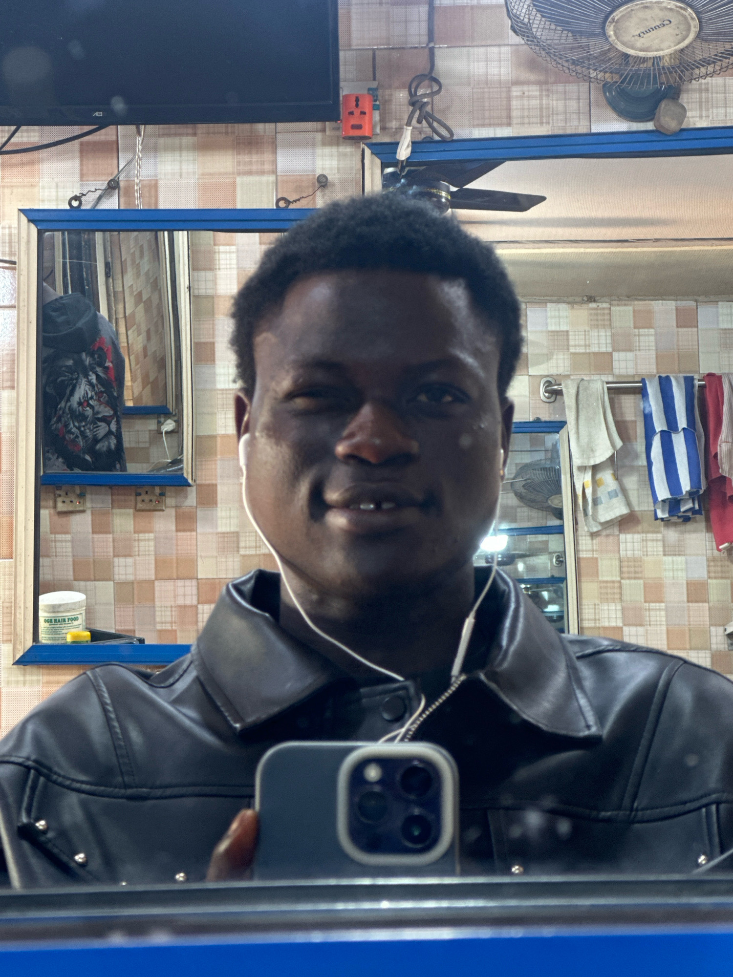 The image is a selfie taken in a barber shop. The subject is a dark-skinned man with short, dark hair, wearing white earbuds and a black leather jacket. He is winking and smiling at the camera, which is held by his reflection in a mirror. The background of the barber shop is visible, showing tiled walls, a shelf with hair products, towels hanging on a rack, an electrical outlet, a ceiling fan, and a television screen. The overall impression is casual and personal, likely taken by the subject himself. The presence of specific barber shop items and the general atmosphere suggest a local establishment in a city like Badagry, Nigeria.