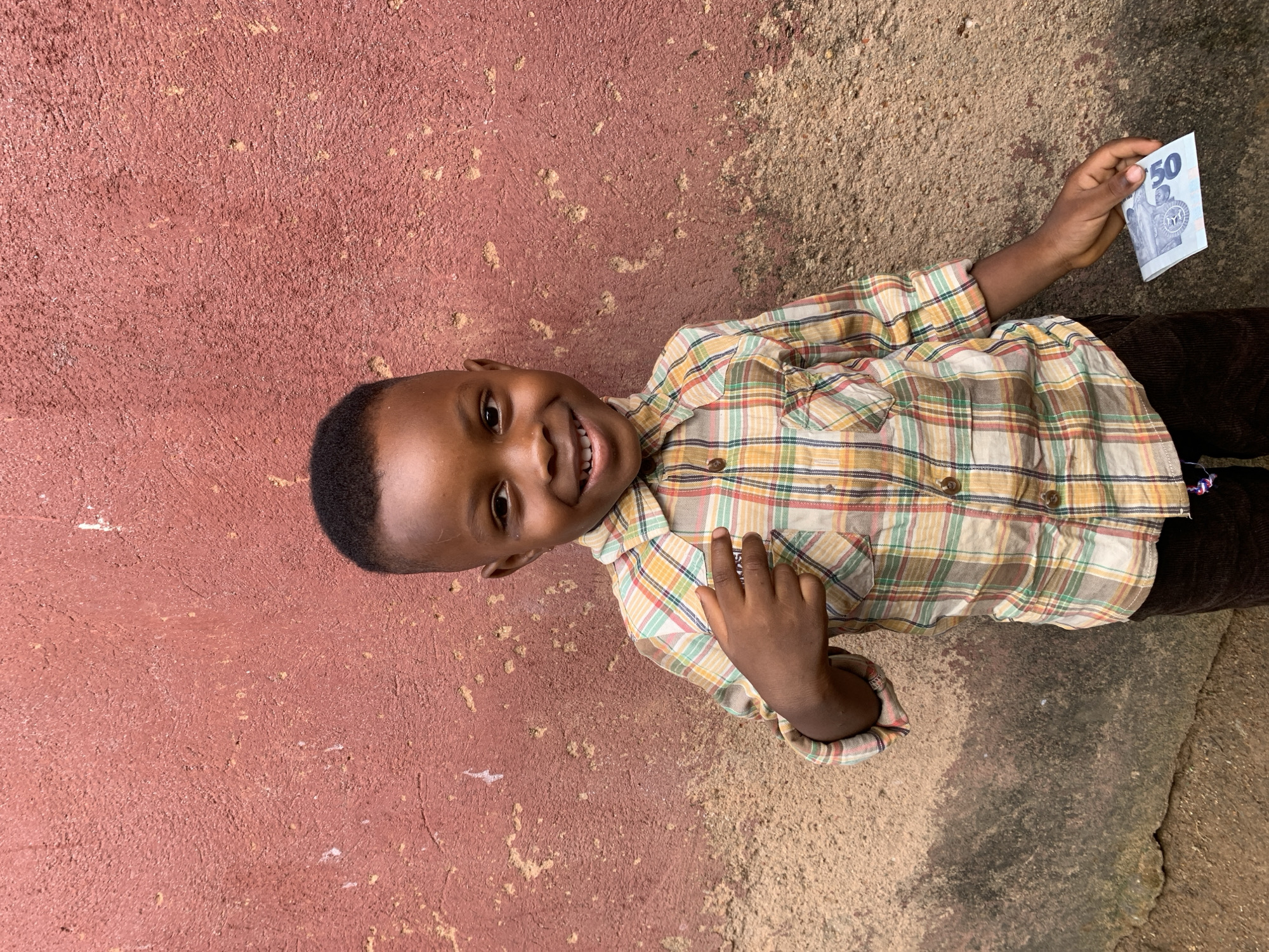 A young child with dark skin and short, neat hair is lying on their side on a surface that transitions from reddish-brown to a more tan, textured ground. The child is wearing a long-sleeved, plaid shirt in shades of yellow, green, and brown, and dark brown pants. They are smiling, facing the camera. In their right hand, they hold a folded banknote with the number "50" prominently displayed. The child appears to be outdoors and the location is Jos, Nigeria.