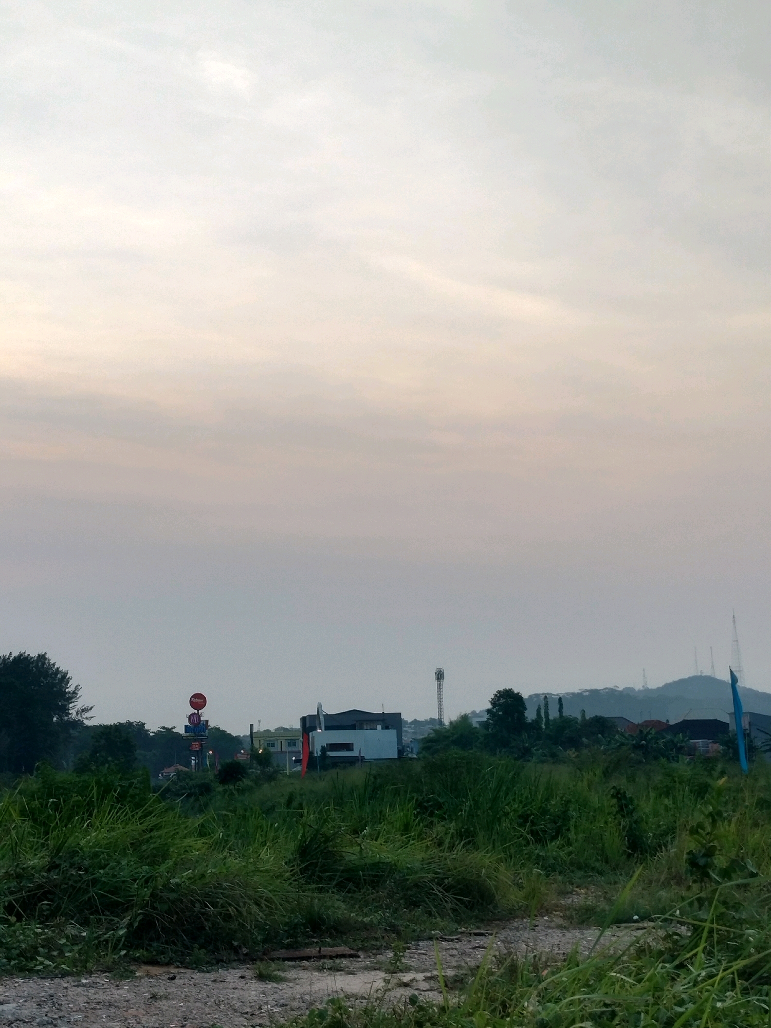 An outdoor, medium shot captures an overcast sky above a landscape in Batam, Indonesia. In the foreground, a gravel path is surrounded by tall, green grass and vegetation. In the middle ground, several buildings are visible, including a white and dark-colored structure with a red flag, a smaller light-colored building, and other structures further back. A tall tower with antennas stands to the left, and more towers are visible on a distant, hazy hill to the right. The scene appears to be a semi-rural area on the outskirts of a city.