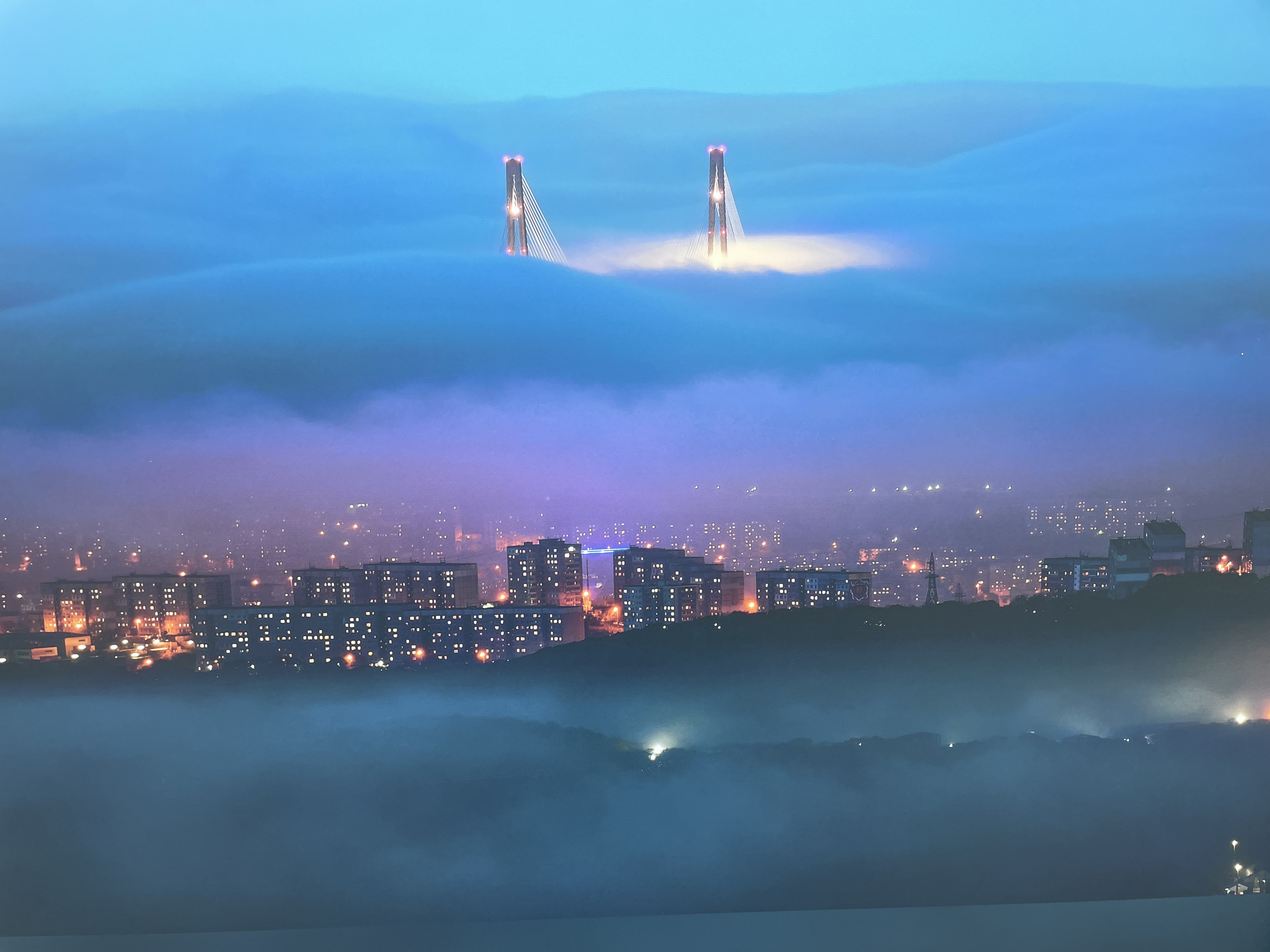 The image presents a city scene at night, likely taken from an elevated perspective, possibly Sochi, Russia. A suspension bridge is partially obscured by dense fog or low clouds, with only the upper portions of its towers visible. The fog creates a surreal effect, as if the bridge is floating above the city. Below the fog layer, a dense urban area is visible, with numerous buildings illuminated by their internal lights. The streetlights add to the overall glow of the cityscape. A second layer of fog obscures the lower part of the city. The sky above the bridge is a soft gradient of blue, indicating either pre-dawn or post-sunset conditions. The absence of discernible people suggests the image captures a still, quiet moment in the urban landscape.