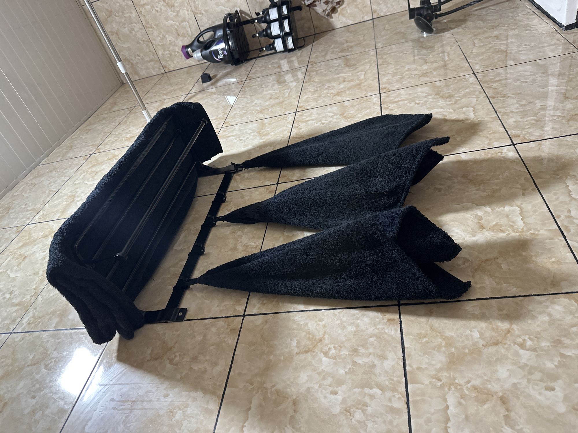 A bathroom rack made of black metal is mounted on a wall tiled with beige marble-patterned tiles. A black towel is folded and placed on the top shelf of the rack. Three black towels are hanging from hooks below the rack. On the right side of the image, there is a black metal shelf with several bottles of toiletries. The ceiling is white and paneled. The image appears to be taken in a bathroom in Fergana City, Uzbekistan, based on the provided metadata. There are no people visible in the image.