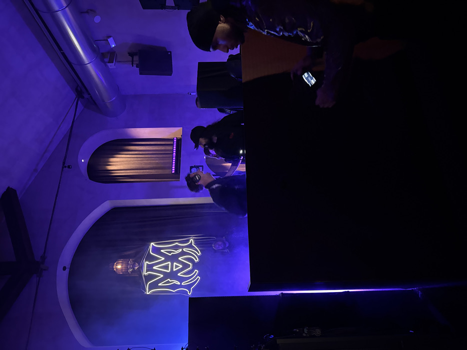 The scene depicts an indoor event venue in Milan, Italy, illuminated predominantly by purple and blue ambient light. A large, white neon sign displaying a stylized "MM" logo, indicative of Marcelo Burlon County of Milan, is prominently displayed on a dark curtain backdrop situated within an arched recess. Above the neon sign, an ornate, dark-colored lantern fixture hangs. Additional architectural features include another arched opening with a dark curtain on the right and a large, exposed metal ventilation duct traversing the upper right ceiling.
In the mid-ground, positioned behind a dark, waist-high structure, likely a DJ booth, two male individuals are actively engaged. The man on the left, with curly hair and headphones resting around his neck, is smiling and looking towards the right. The man on the right, wearing a dark baseball cap and possessing long dark hair, holds a microphone to his mouth, appearing to perform or address the audience. A third, less distinct figure is faintly visible in the background's purple haze.
In the immediate foreground, partially obscured on the right, a man wearing a dark beanie and a dark, patterned jacket is depicted in profile, intently viewing and interacting with a smartphone held in both hands. The overall environment suggests an active social gathering or performance event.