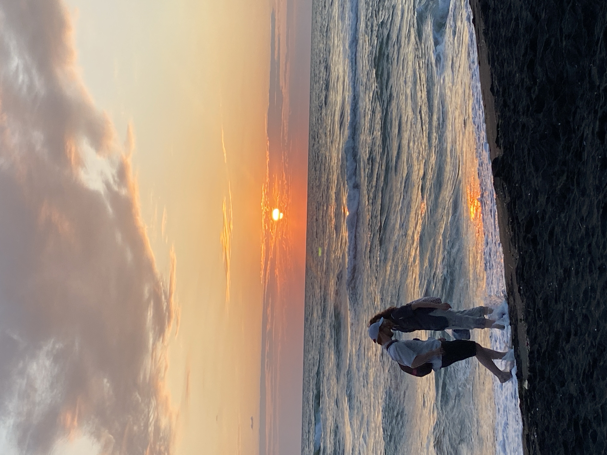 The image captures a romantic scene on the black sand beach of Ureki, Georgia, during sunset. The sky is painted with hues of orange, pink, and yellow, with scattered clouds tinged by the setting sun. The ocean, with its moderate waves, reflects the warm colors of the sky, creating a shimmering effect. In the foreground, a couple stands close together at the water's edge, their faces obscured as they share a kiss. They are both dressed casually, with the male figure wearing a light-colored t-shirt and shorts, and the female figure in darker attire. Both have backpacks, suggesting they may be travelers. The dark, wet sand contrasts with the foamy waves washing over their feet, highlighting the moment of intimacy against the backdrop of the vast sea and sky.