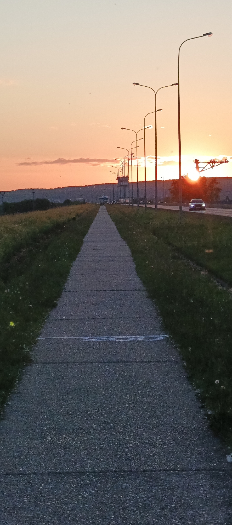 This image captures a sunset scene along a pathway and road in Chaikovsky, Russia. A concrete pedestrian pathway, segmented by horizontal lines, leads the viewer into the frame, bordered by grassy verges. Along the right side of the pathway, a paved road stretches into the distance, with streetlights lining its edge. Several cars are visible on the road, some moving towards the viewer and others away. The sky is dominated by the warm hues of a sunset, with soft orange and pink clouds and the sun itself peeking through the trees on the horizon, casting a bright glow. In the background, a low, tree-covered hill forms the distant landscape.