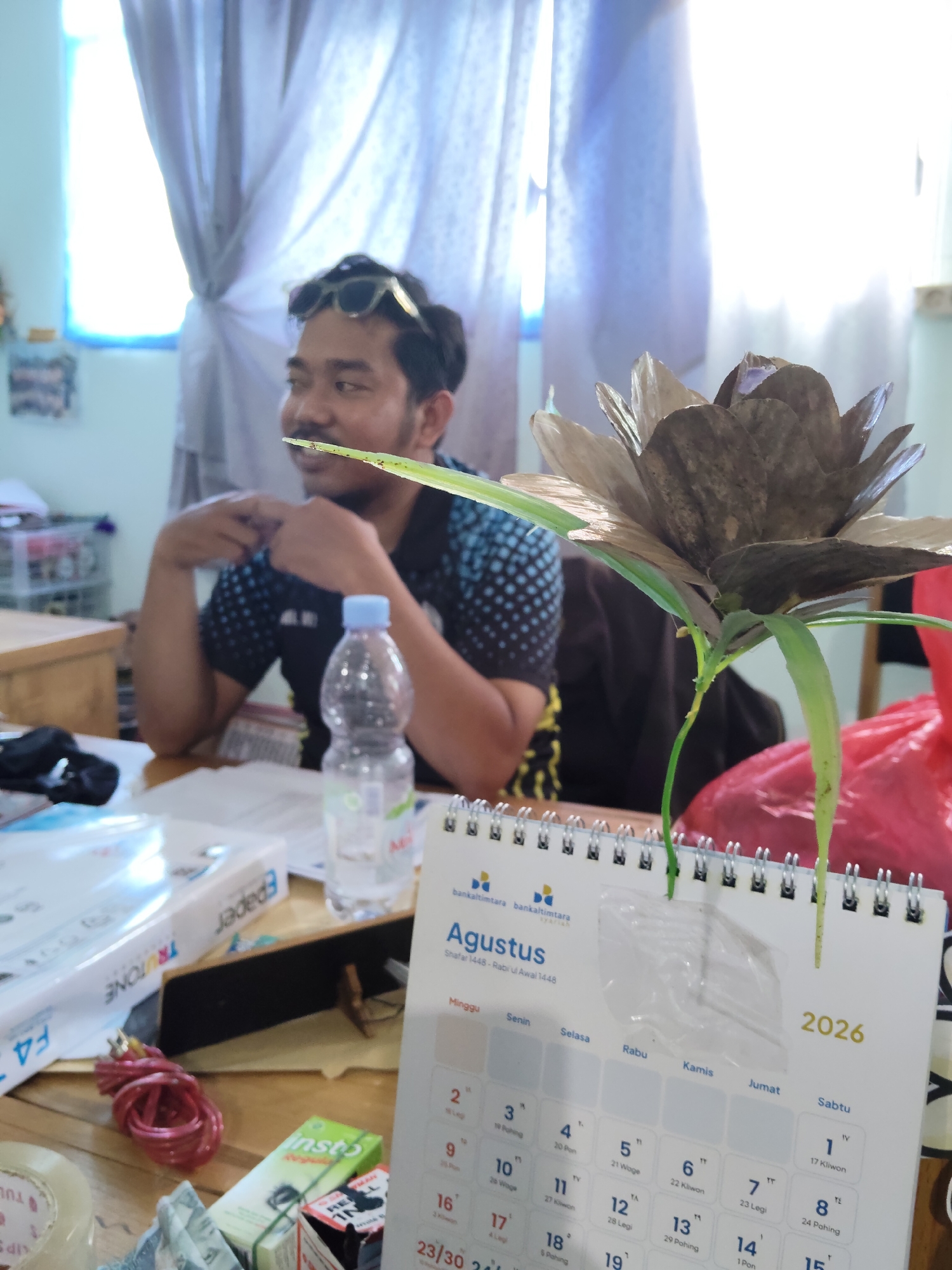 The image depicts a man in a black and blue patterned shirt sitting at a wooden desk with various items scattered around him. He is wearing sunglasses on top of his head and looking to his left. In the foreground, a calendar for August 2026 is open, showing the days of the week and dates. A plastic bottle of water, a roll of red wire, a package labeled "insto," and a box of papers are also visible on the desk. A decorative plant with brown petals and green leaves is placed in front of the calendar. The background shows a light blue curtain and a window. The scene appears to be an indoor workspace, possibly an office or a home study, in Lori, Indonesia.