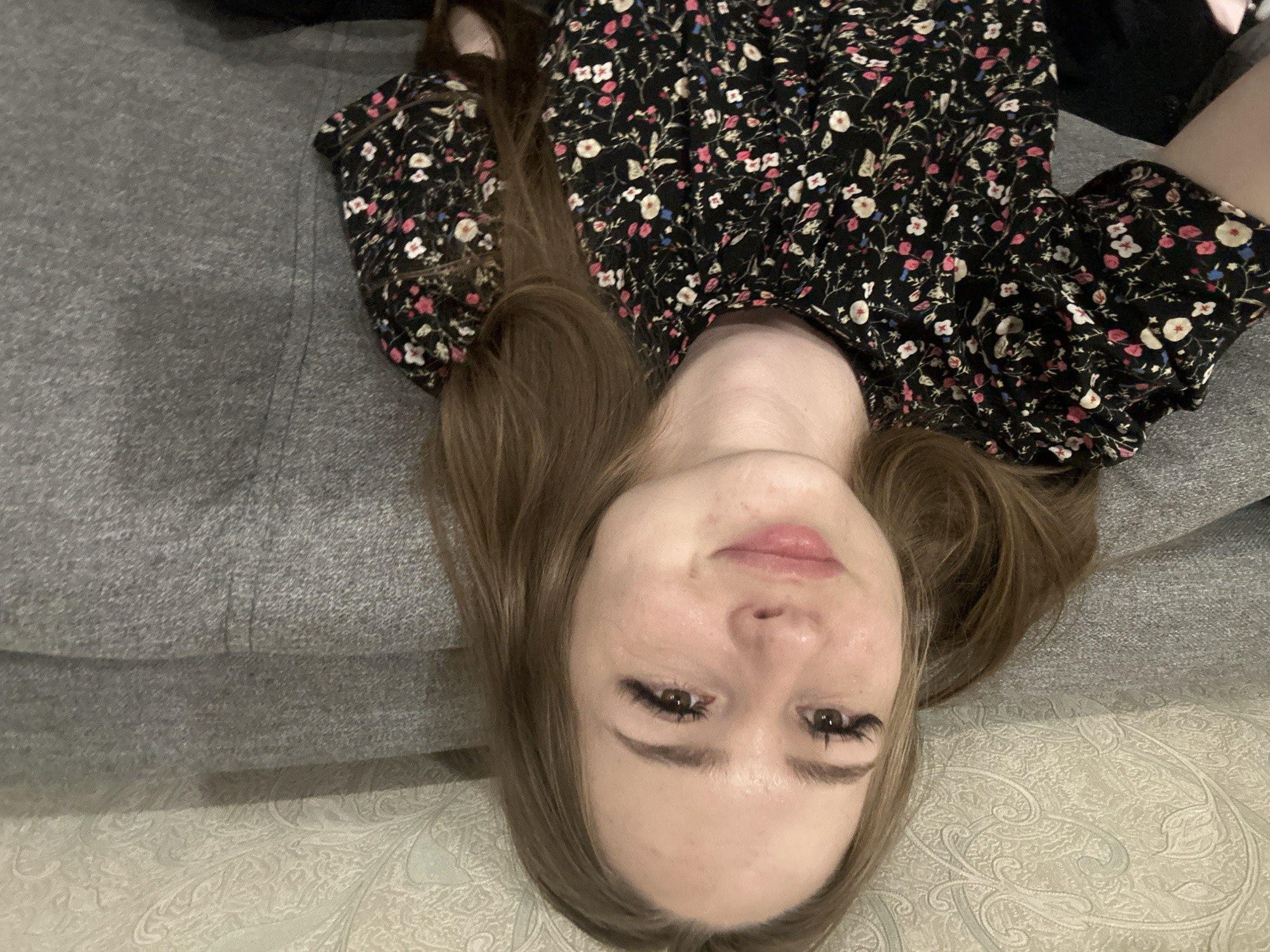 A young Caucasian woman with light brown hair pulled into two pigtails is looking at the camera with a slight smile. She is wearing a black short-sleeved dress with a floral pattern. She is sitting on a grey sofa, with a textured wallpapered wall behind her. The lighting appears to be artificial, possibly from an overhead source, casting a slight shadow on the wall behind the woman. The overall setting suggests an indoor environment, possibly a living room.