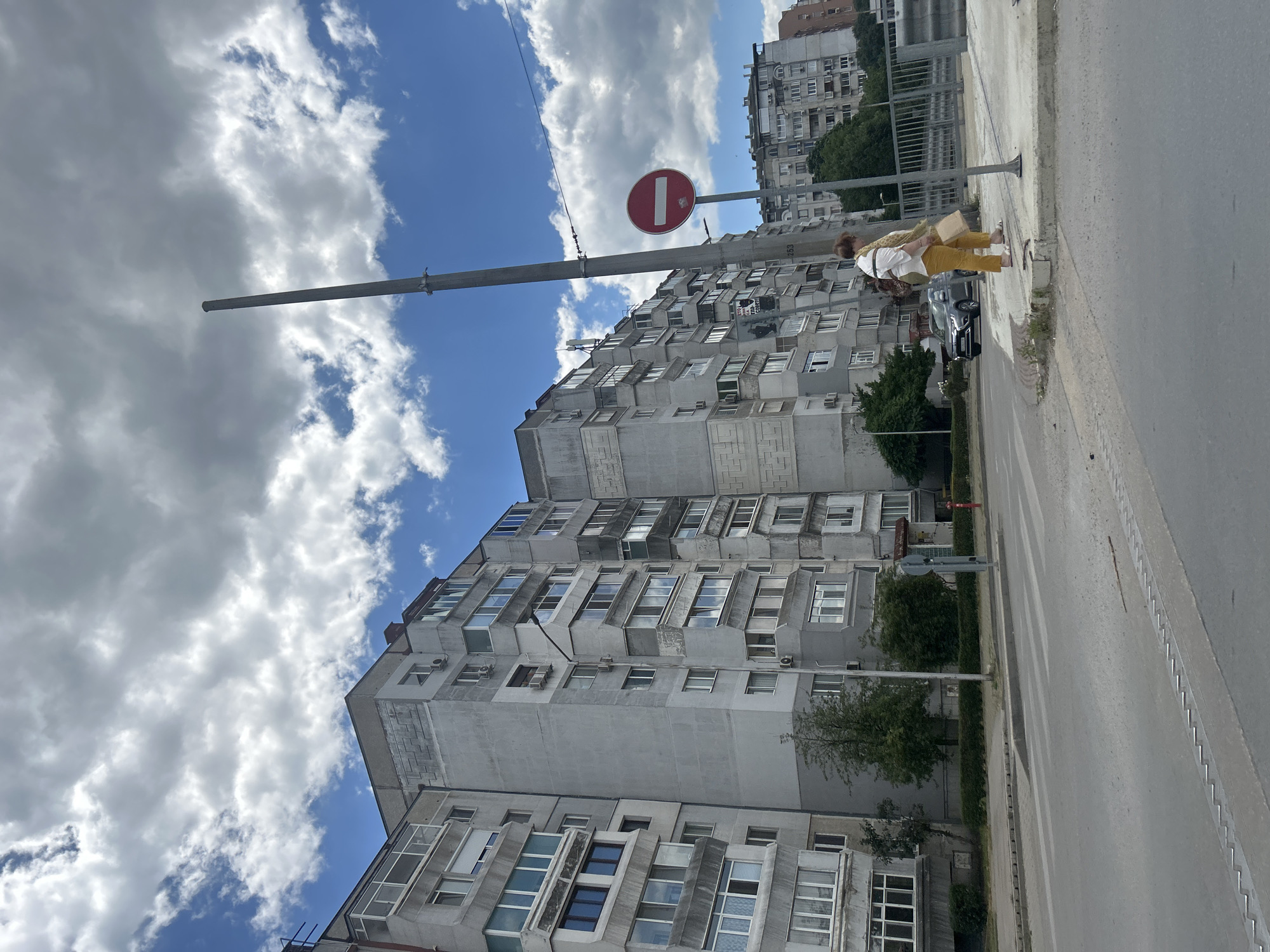 The image depicts a street scene in Varna, Bulgaria. A row of identical, grey, Soviet-era apartment buildings dominates the left side of the frame. These buildings are characterized by numerous windows, balconies, and visible air conditioning units. In the foreground, a paved road with pedestrian crossings is visible. On the right side of the street, a woman in a white shirt and yellow pants stands on a sidewalk, holding a shopping bag. She is near a tall metal pole with a red and white circular "no entry" sign attached. A grey car is parked on the street to the left of the woman. The sky is partly cloudy with dramatic white and grey clouds against a bright blue sky. In the background, more apartment buildings and trees are visible. The overall impression is of a typical urban residential area on a cloudy day.