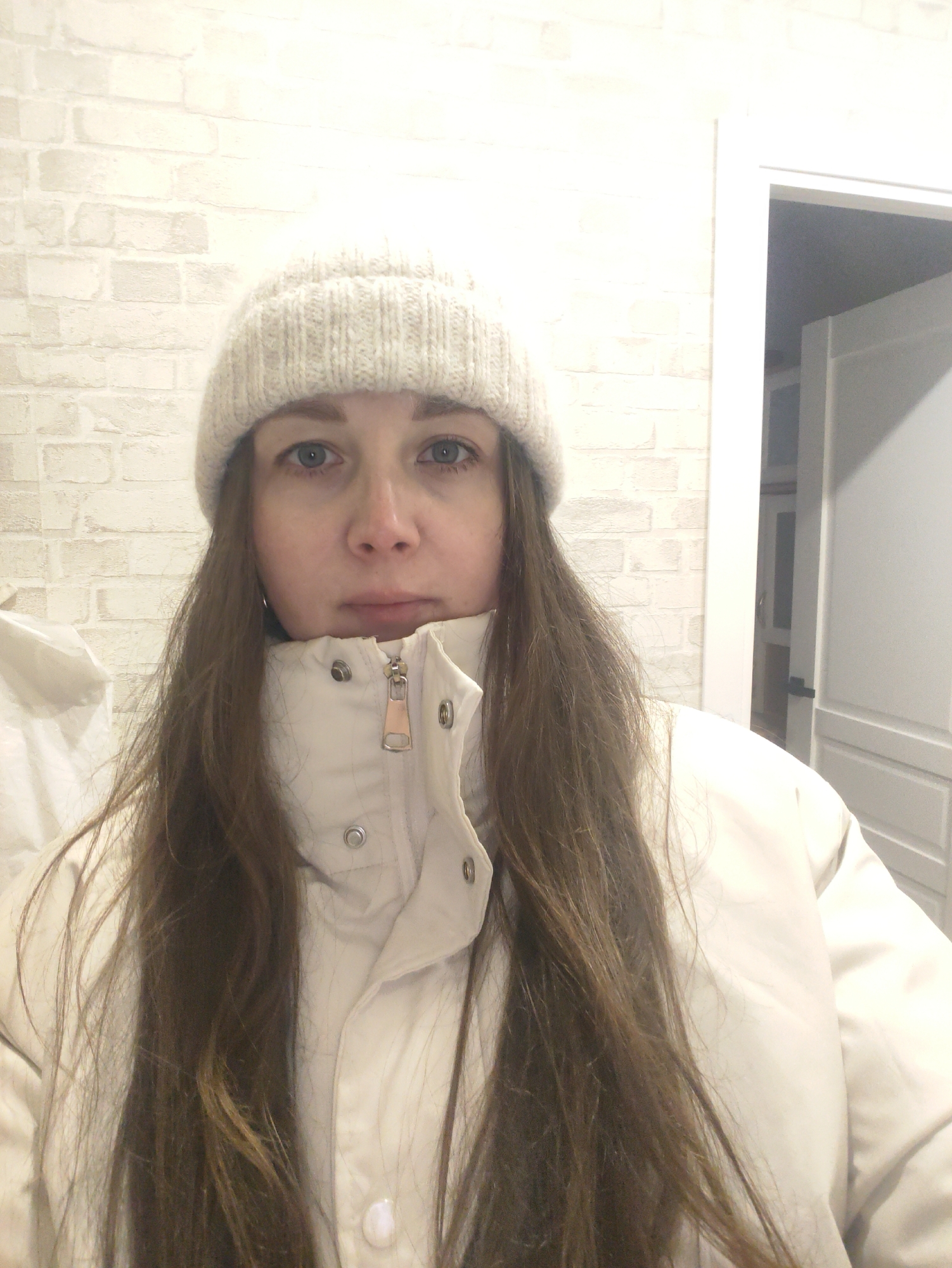 A woman is centered in a medium shot. She is wearing a light-colored knitted hat, a light-colored puffer jacket with a zipper and buttons, and has long brown hair. She faces the camera directly. The background is a white brick wall and a partially visible white doorframe with an open white door. The location is in the city of Unknown, Unknown.