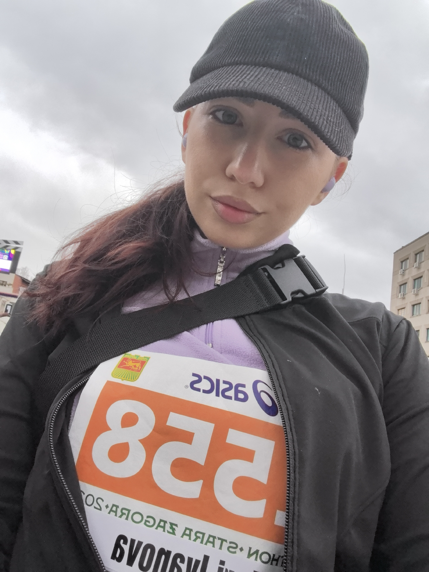 A selfie of a young woman wearing a black corduroy cap, lavender-colored jacket, and a black running jacket, with a bib number "258" attached to the front. She has light skin, light-colored eyes, and her dark hair is pulled back in a ponytail. She has wireless earbuds in her ears. The bib has an orange rectangle with the number "258" written in white. The Asics logo is visible at the top right corner of the bib. At the bottom of the bib is text written in Cyrillic letters. She also wears a black cross-body bag.
The background shows a cloudy sky and buildings. From the style of the building and the Cyrillic text on the bib, it can be determined that the location is Stara Zagora, Bulgaria.