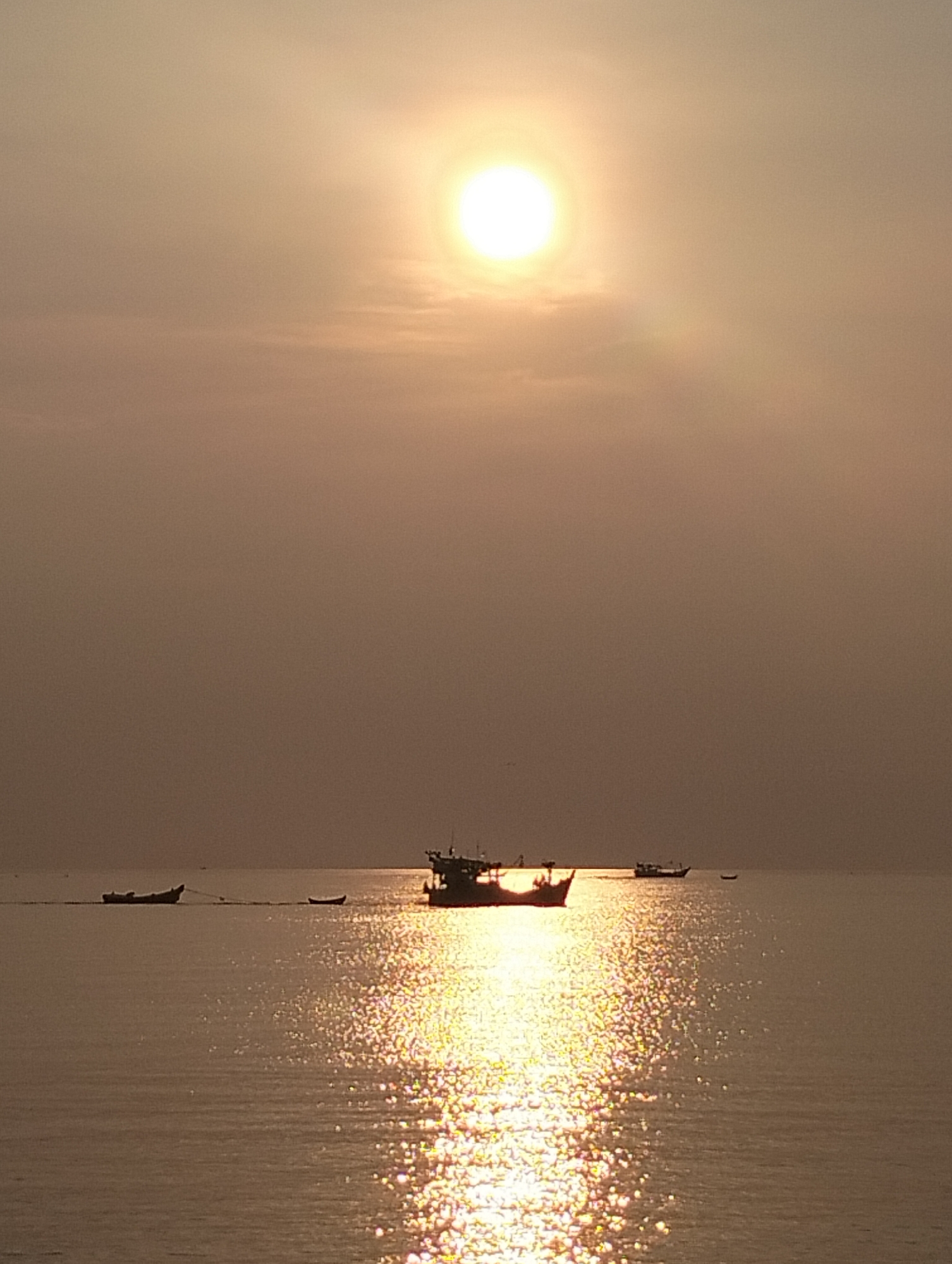 The image captures a seascape at sunset, likely in Lhokseumawe, Indonesia, given the prompt. The sun, a bright disc, is high in the sky, its light filtered by the atmosphere, creating a hazy, golden effect. The sea, which covers the lower portion of the frame, reflects the sun's light in a shimmering, glittering pattern. Several fishing boats are silhouetted against the bright water. One larger boat is more prominently placed near the center, with smaller vessels to the left and further in the distance to the right. A small boat is being towed behind the left-most vessel. The sky is a gradient of golden hues, darkest at the top and lightening towards the horizon. No people are visible in the image.