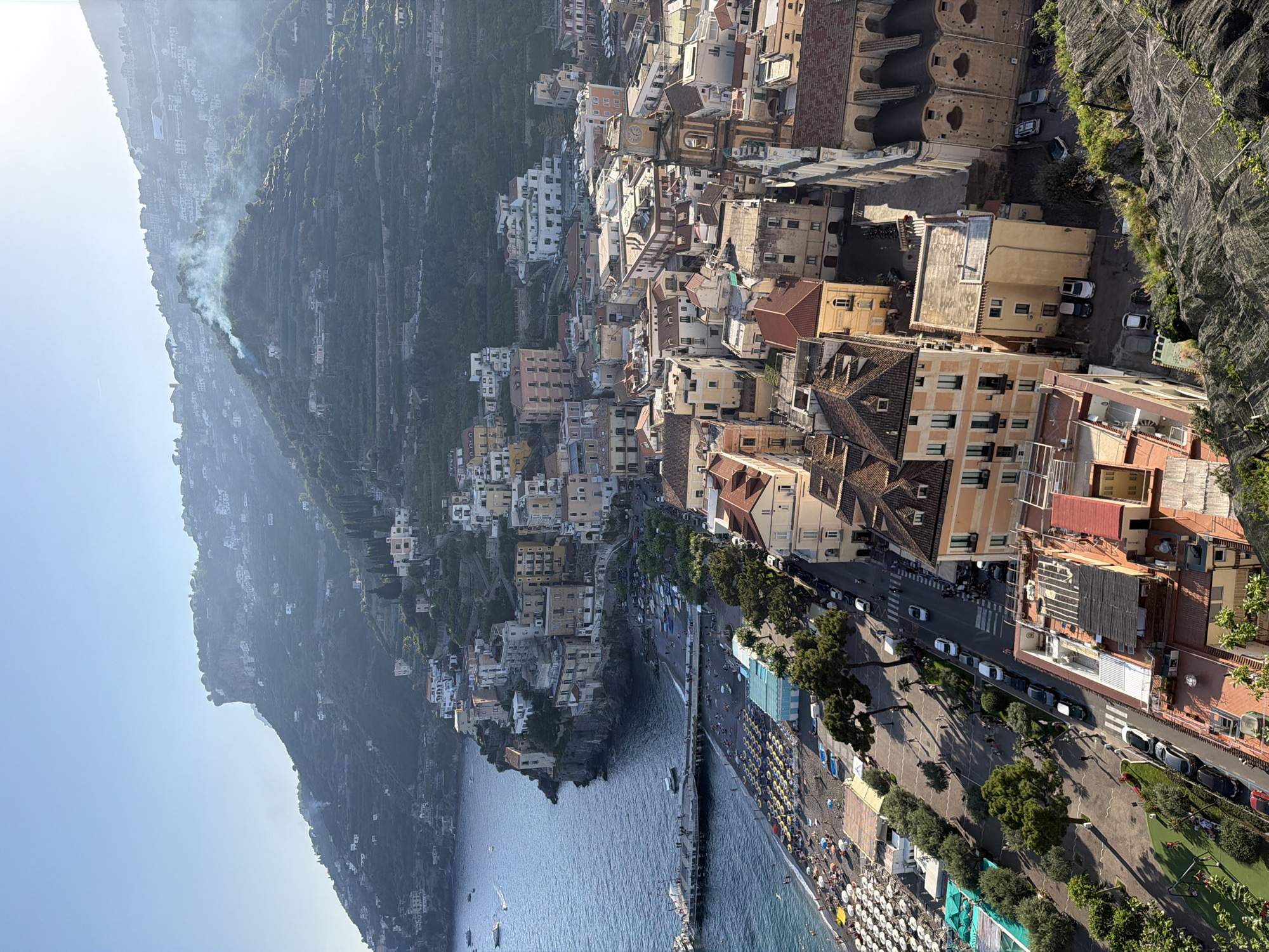 The image presents a high-angle view of Minori, Italy, with the Tyrrhenian Sea prominently featured. A pier extends into the sea, and a beach is visible, densely populated with sunbathers and umbrellas. The coastline is lined with trees and a road with parked cars. Buildings are clustered together, ascending the hillside, showcasing diverse architectural styles and colors. The background features a steep, terraced mountain, possibly used for agriculture, with some smoke rising from it. The sky is clear blue.