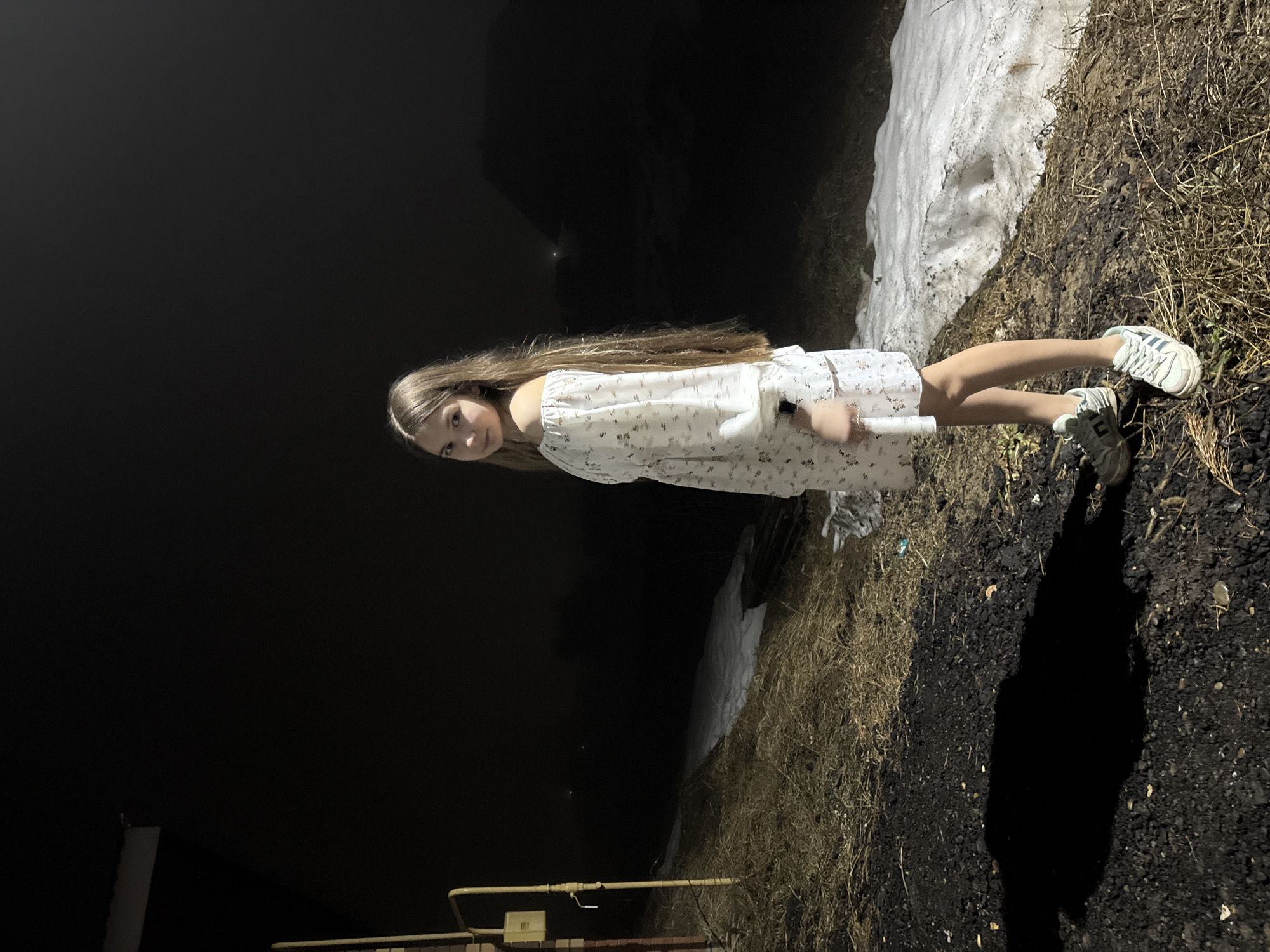 The image shows a young girl standing outside at night in Garevaya, Russia. She is wearing a light-colored dress with floral patterns and white sneakers. She is holding a cylindrical object covered in a white cloth. To her right is a patch of ground covered with dark soil and dry grass, transitioning to a snow-covered bank. The background is dark, suggesting it is night, with a few faint lights visible in the distance. A gas line is partially visible in the lower left corner. The girl appears to be posing for the photo.
