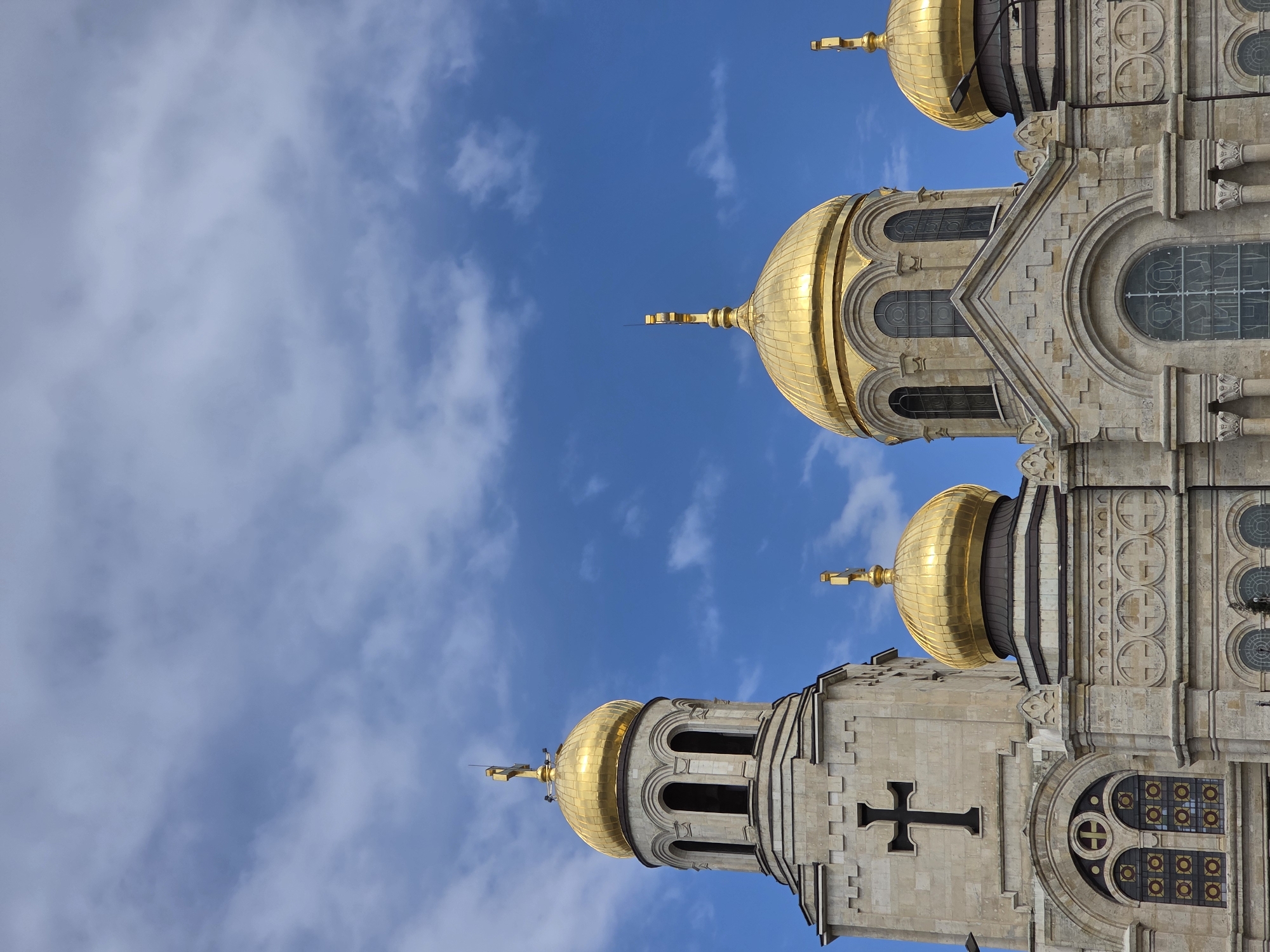 The image shows the domes and upper structure of the Dormition of the Mother of God Cathedral in Varna, Bulgaria. The cathedral's architectural details include gold domes, ornate stone carvings featuring crosses, arched windows, and decorative elements on the exterior walls. The domes are gleaming against a blue sky with scattered white clouds. The composition is from a low angle, emphasizing the cathedral's height.