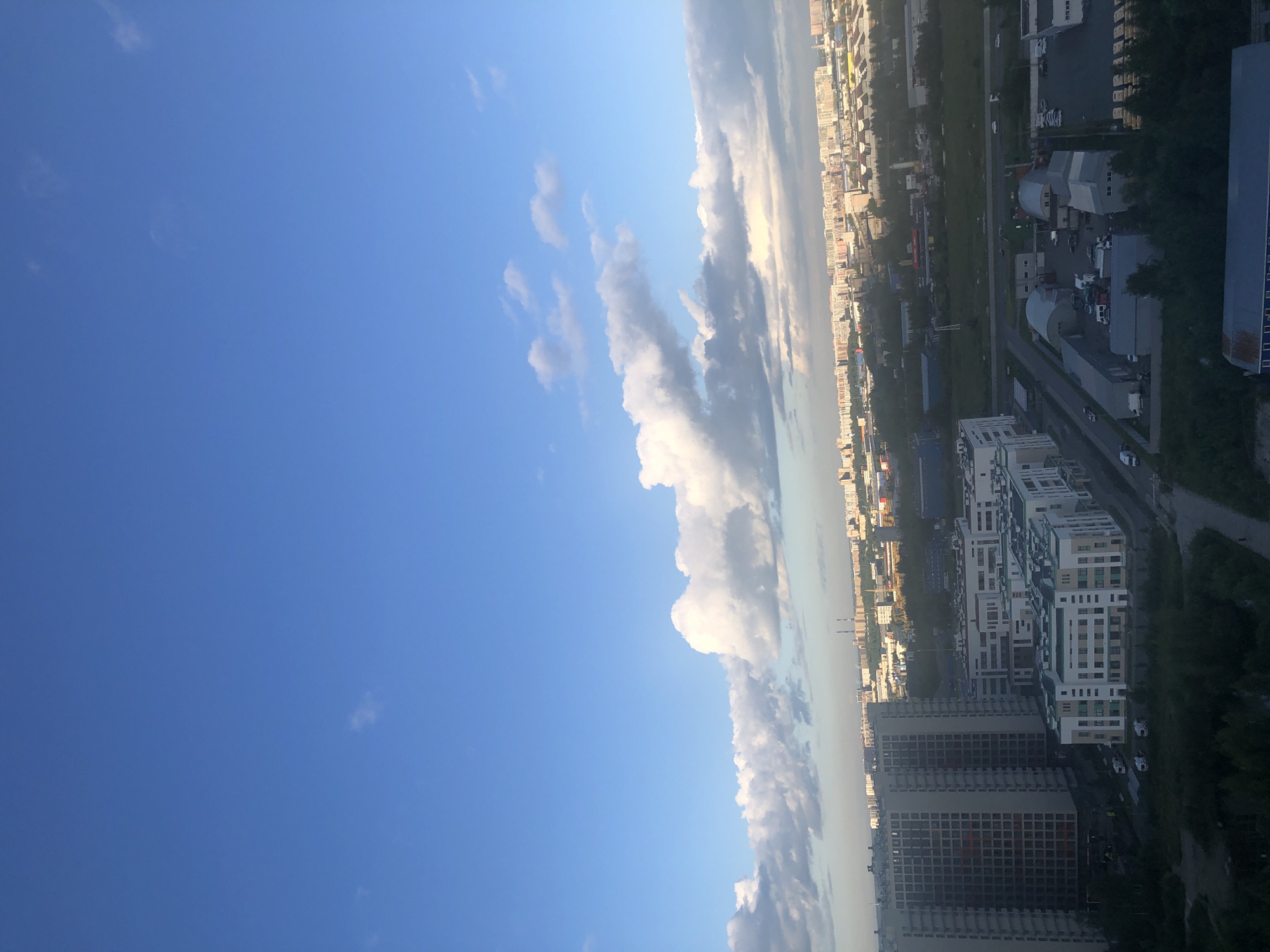 A high-angle, wide shot captures a city skyline under a bright blue sky with scattered white clouds. The foreground features several modern apartment buildings, one of which is unfinished with visible concrete and scaffolding. Adjacent to the apartment buildings are lower-rise structures with white facades and green accents, possibly a school or office complex, and a parking lot with several vehicles. Further into the midground, industrial buildings with curved roofs and an open yard filled with trucks and shipping containers are visible. The background is dominated by a dense urban landscape of numerous multi-story buildings stretching to the horizon. Trees line the bottom of the frame in the foreground, and a road with a few moving cars can be seen. The lighting suggests it is daytime, likely late afternoon or early evening due to the long shadows cast by some of the buildings.