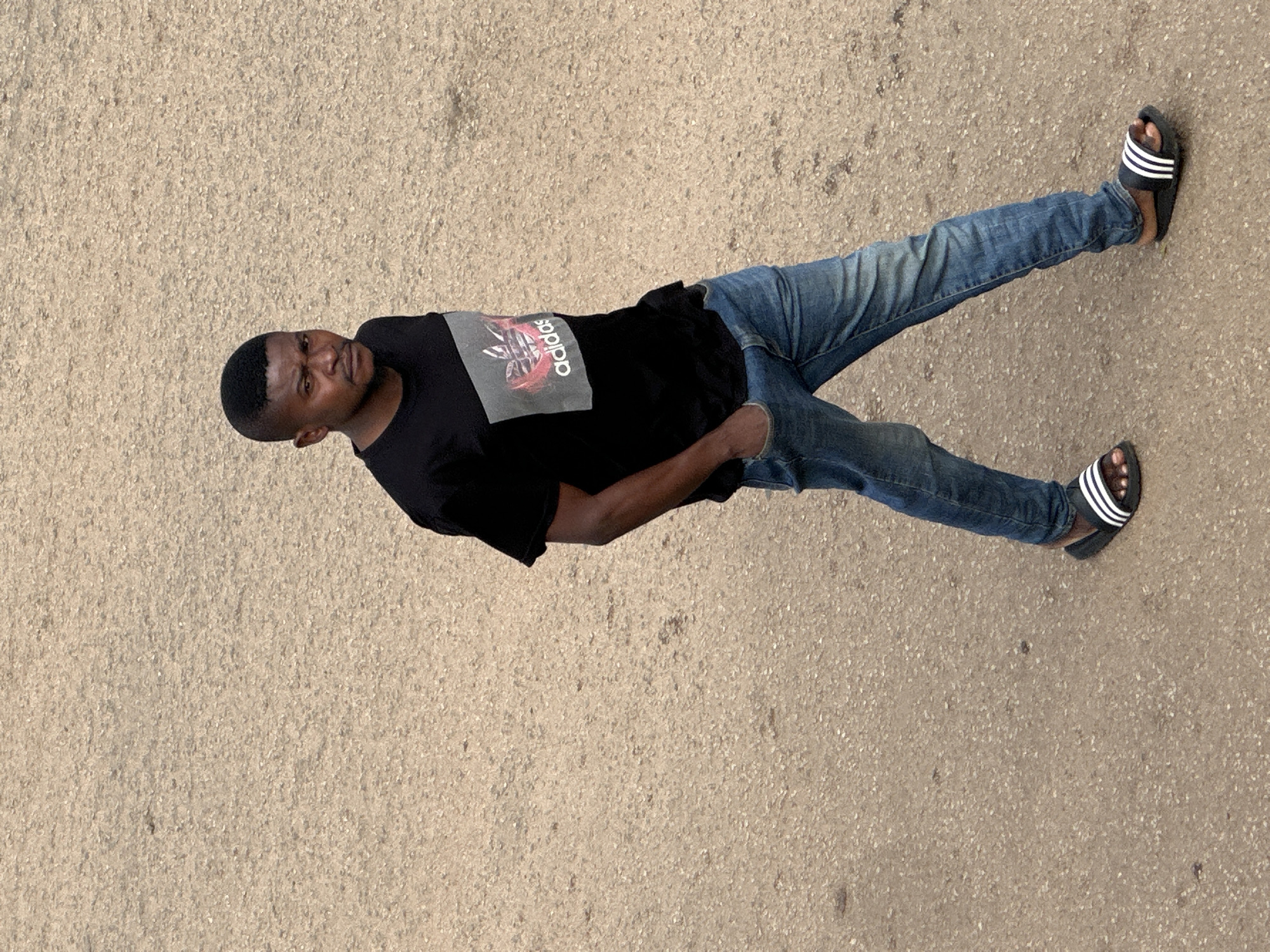 In Bukuru, Nigeria, a young Black man is captured in a full-body shot from a high angle, walking on a textured, sandy-colored ground. He is wearing a black t-shirt with a graphic Adidas logo in shades of pink and grey, and faded blue jeans. His right hand is tucked into his jeans pocket, and he is wearing black Adidas slides with white stripes. The man is looking towards the camera with a serious expression. The background is an expanse of the same textured ground, indicating an outdoor, urban setting.