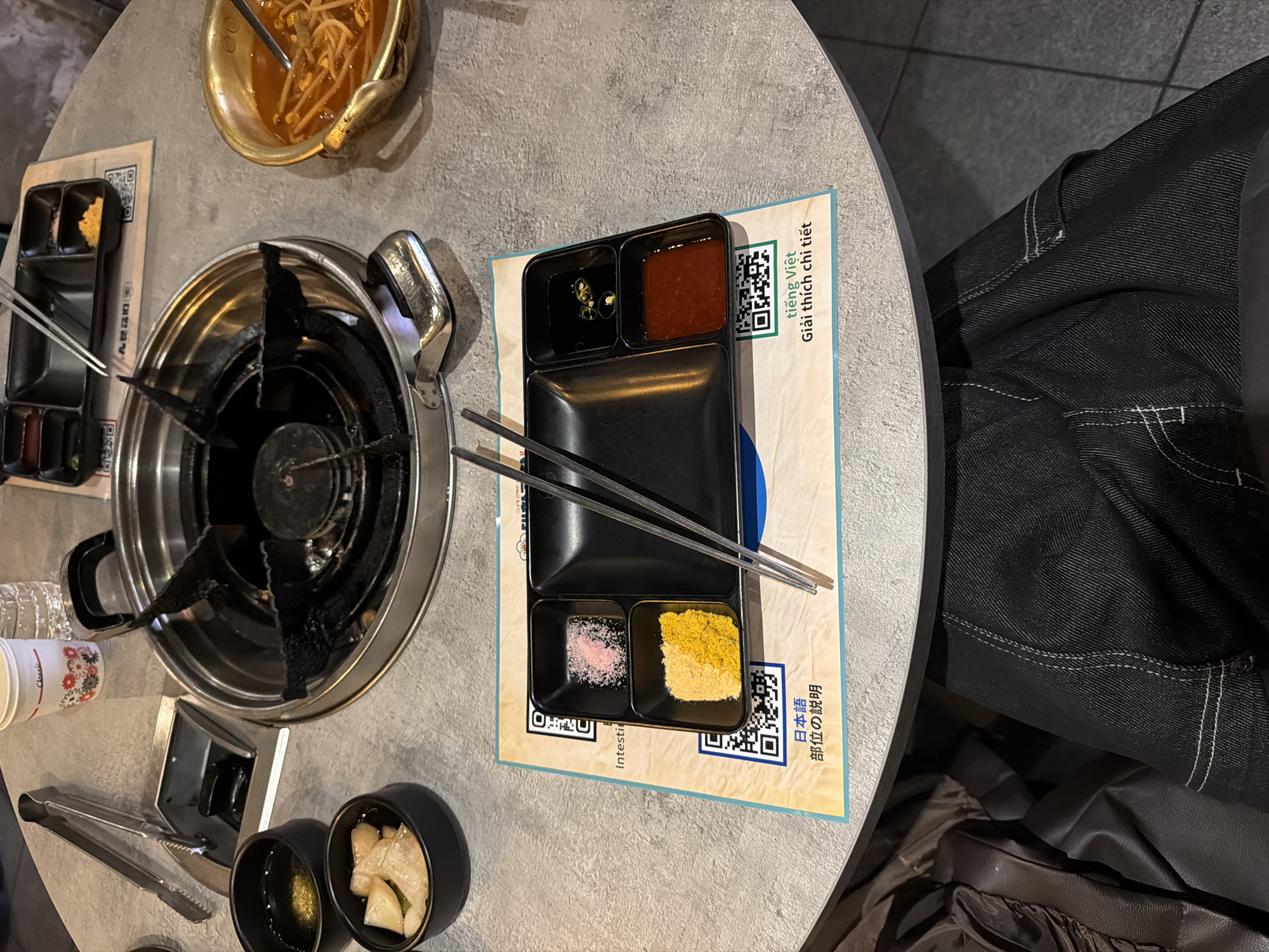 Here's a detailed description of the image:
**Scene:** The image appears to be a top-down view of a Korean BBQ setting, likely inside a restaurant.
**Location:** Given the use of Vietnamese and Japanese languages on the placemat (along with the Korean BBQ setup), it is likely in an ethnically diverse area, such as Seoul, South Korea, where international cuisine and languages are prevalent.
**Objects:**
* A built-in gas burner for grilling.
* A pot of what appears to be bean sprouts.
* Small bowls containing sauces/dips and pickled radish.
* A divided dish with different condiments (possibly soy sauce with chili slices, a red pepper paste, pink salt, and seasoned salt).
* Chopsticks.
* Tongs.
* Disposable cups.
* Placemats with Vietnamese and Japanese text, likely providing explanations of the Korean BBQ experience.
* Dark denim jeans, suggesting a person is seated at the table.
**Possible Actions/Interactions:**
* Preparing to cook or eat Korean BBQ.
* The person is likely dining alone or with others out of frame.
**Environment:**
* The table is round with a grey surface.
* The flooring is tiled.
* The scene is indoors, presumably a restaurant.