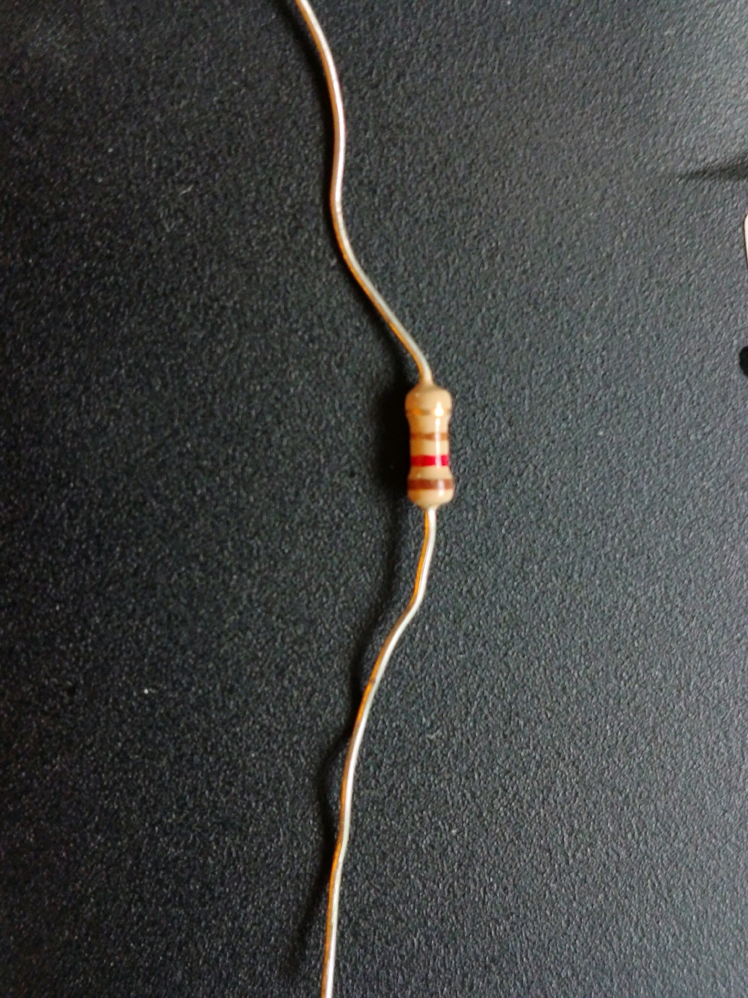 A single resistor lies on a black textured surface. The resistor is cylindrical with a light brown body and three colored bands: a wider brown band followed by a thinner red band, and then another wider brown band. Two thin wires extend from either end of the resistor, curving away from it. The surface appears to be a table or countertop.