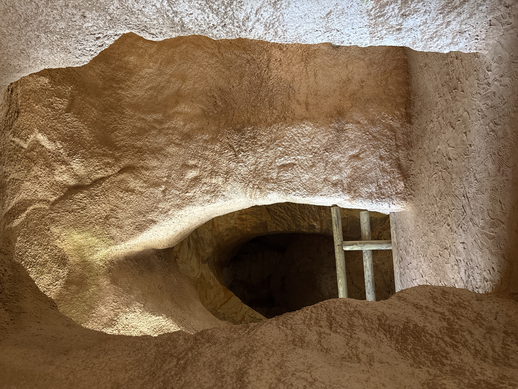 The image depicts the interior of a sandstone cave in Piusa, Estonia. The cave has natural, rough-hewn walls with visible layers of sandstone. In the foreground, the cave opens into a sandy pathway marked with numerous footprints. A wooden railing, consisting of posts and horizontal bars, is visible, separating the pathway from a darker, deeper section of the cave beyond. The lighting suggests natural light filtering into the cave from an opening, casting shadows and highlighting the texture of the sandstone. The overall scene conveys a sense of natural formation and human passage.