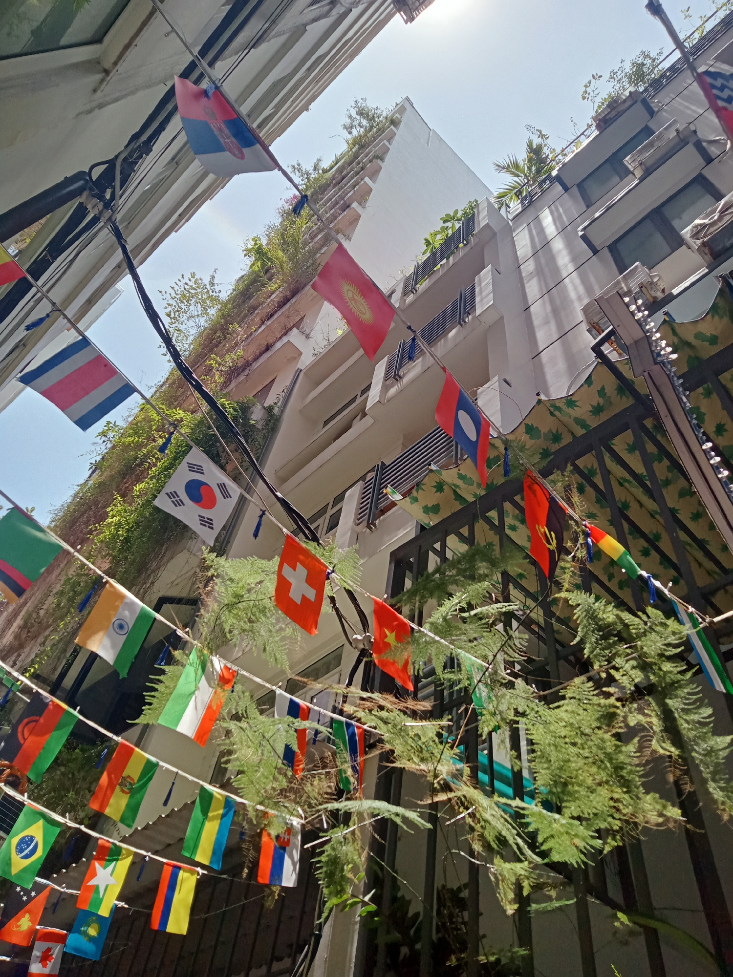 The image shows a view looking up at buildings in Nha Trang, Vietnam, decorated with numerous national flags strung across the narrow space between the buildings. The buildings are pale in color, with vertical lines and some greenery growing on the walls and balconies. The flags represent various nations and are attached to strings that run from one building to the other. A decorative awning is visible on the right side of the frame. The sky is light blue, and the sun is visible above the buildings.
