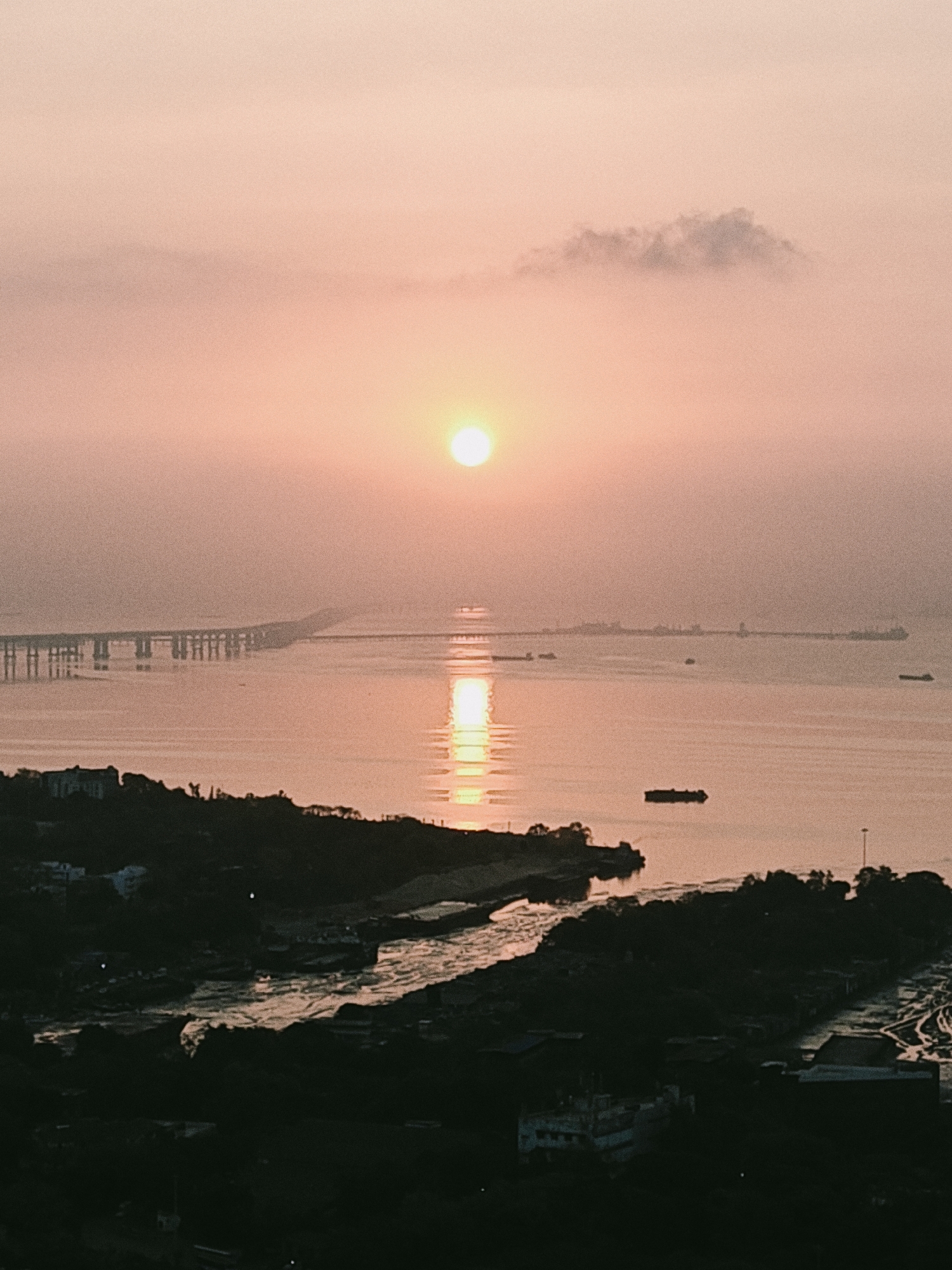 The image captures a sunrise scene in Mumbai, India, featuring the Bandra-Worli Sea Link bridge extending across the Mahim Bay towards the horizon. The sun is positioned centrally in the frame, reflecting a bright vertical streak of light on the water's surface. A light fog or haze permeates the atmosphere, muting colors and obscuring details. Several boats are visible on the water, and in the foreground, the coastline is characterized by dark landmass with trees and urban structures.