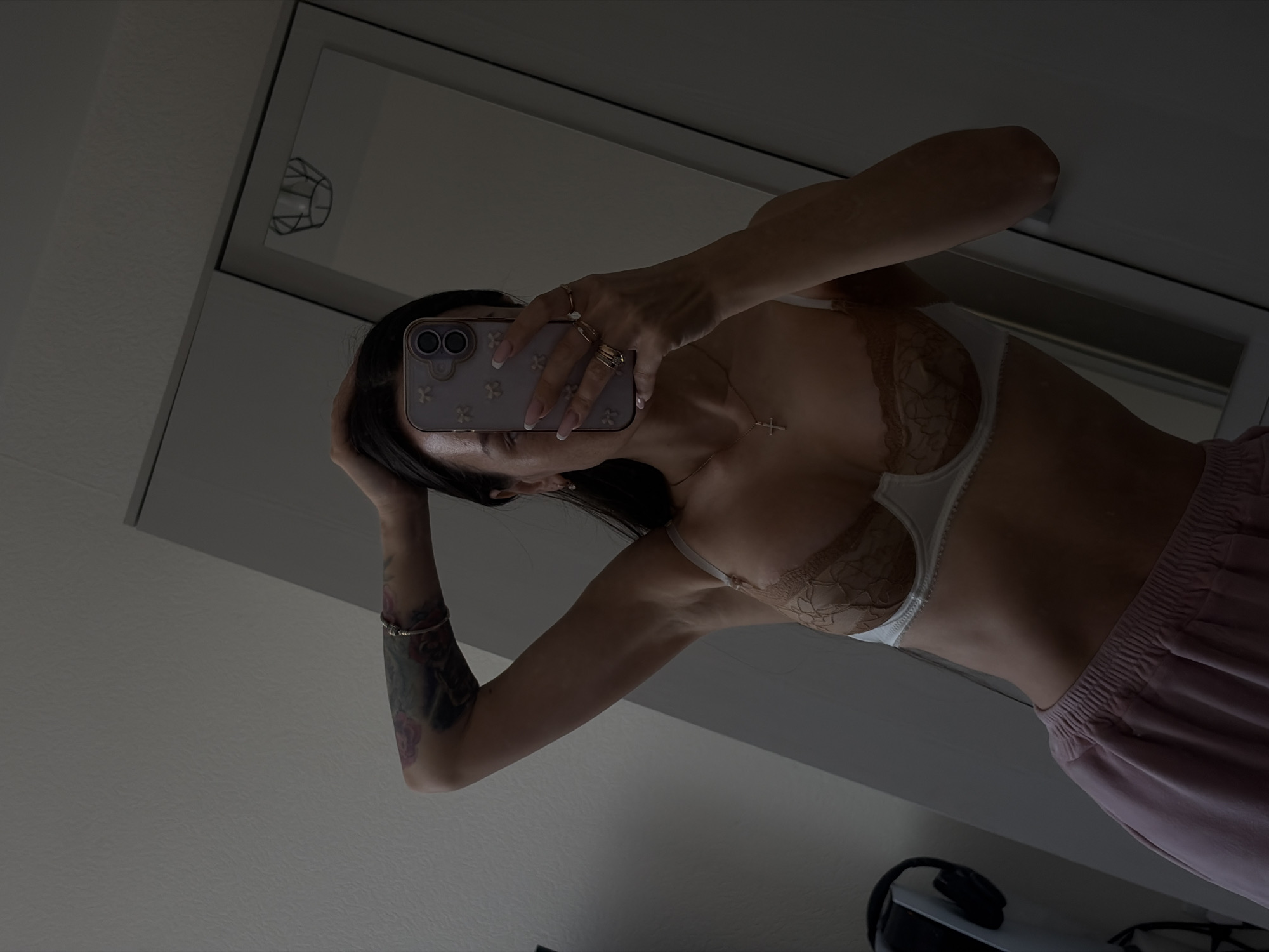 A selfie taken in a room reveals a woman with dark hair, clad in a bra, and light pink pants. She holds a phone with a floral case, obscuring her face. The scene is reflected in a mirror, which partially reveals a light fixture and the room's interior. The woman's left arm displays a tattoo and a bracelet. The setting appears to be indoors, with neutral wall colors.