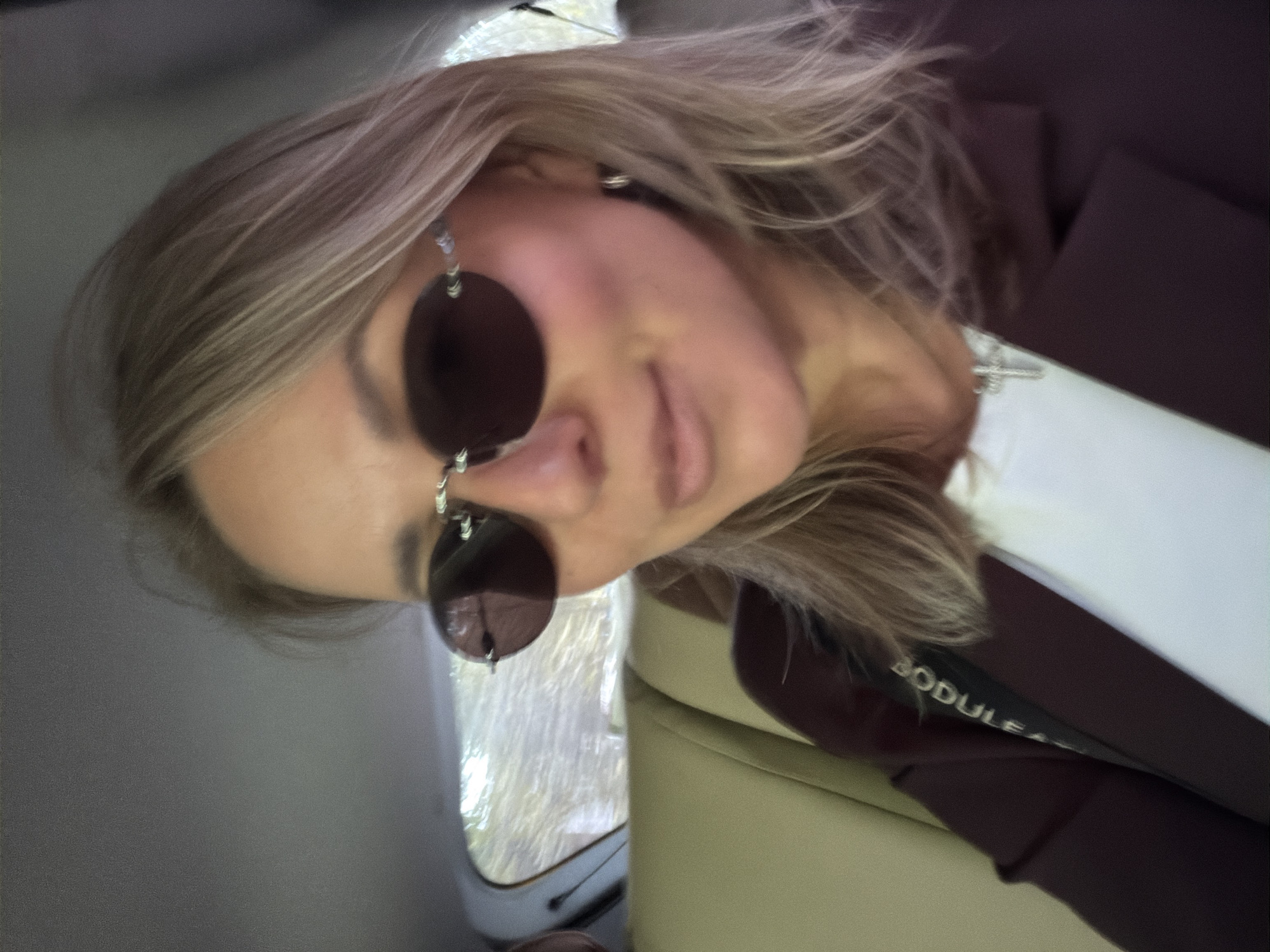 A woman is photographed indoors, likely in a vehicle given the window on the left side, with a cream-colored leather-like seat visible in the lower right. She is wearing sunglasses with circular lenses and a burgundy blazer over what appears to be a white top. The label "BODULEA" is partially visible on the blazer. Her hair is blonde and styled in loose waves. The location is unknown.