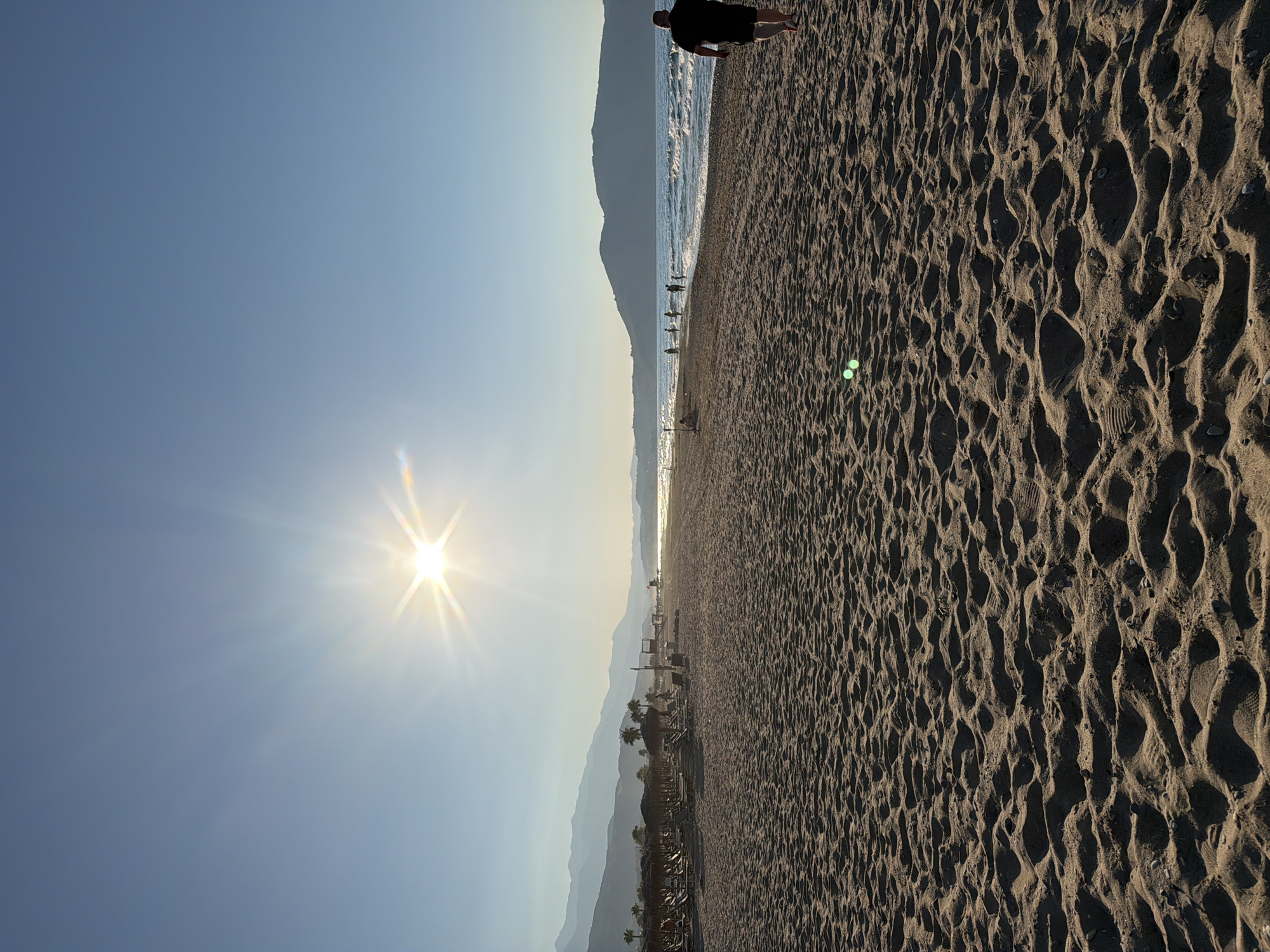 The image depicts a wide, sandy beach in Dramia, Greece, bathed in the warm glow of a setting sun. The sky is a clear, serene blue, with the sun's rays creating a starburst effect. In the foreground, the textured sand is imprinted with numerous footprints, suggesting recent activity. A lone man, wearing a black t-shirt and shorts, walks away from the camera towards the right side of the frame.
In the mid-ground, the beach transitions to a more populated area with rows of straw umbrellas and lounge chairs, indicative of a resort or tourist area. Several people are visible scattered along the shoreline, some wading in the gentle waves of the sea, while others are walking along the beach. The sea is calm with small waves breaking on the shore.
In the background, a chain of mountains rises majestically, their peaks softened by the hazy atmosphere of dusk. The overall scene conveys a sense of tranquility and the peaceful end of a day at the beach in Dramia, Greece.