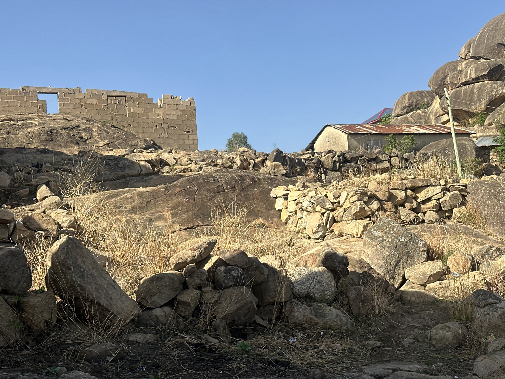 The scene depicts a rocky landscape in Jos, Nigeria. In the foreground, a collection of rocks and dry vegetation dominate. An unfinished brick structure with window openings is visible on the left side, situated atop a large rock formation. To the right, a small building with a rusty corrugated metal roof is nestled amongst the rocks and boulders. A stone wall partially obscures the building. The sky is clear and blue. A utility pole is also visible on the right.