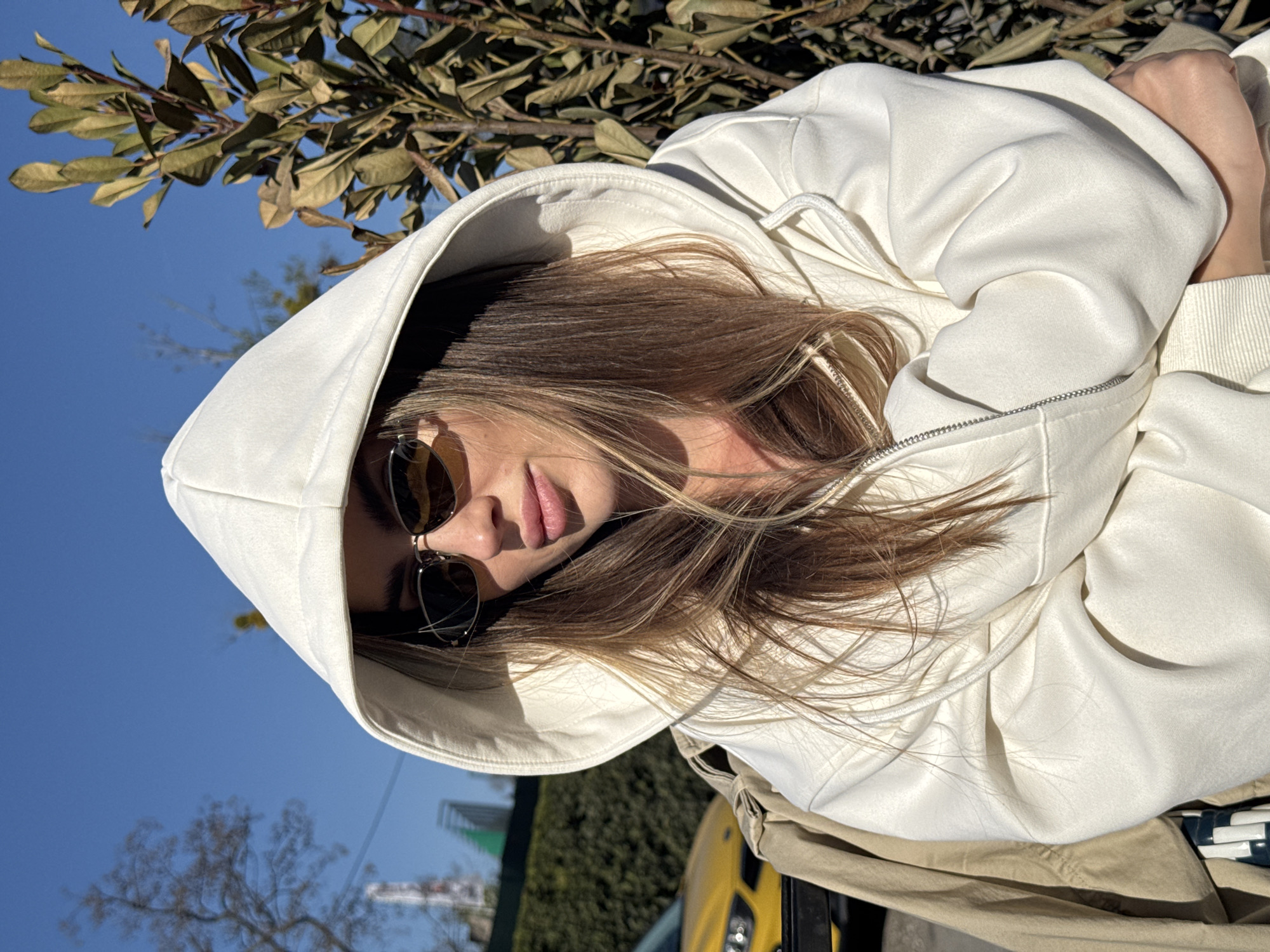 The image presents a Dutch-angled medium close-up portrait of a person, likely female, captured outdoors on a clear, sunny day in Warsaw, Poland. The subject is seen from a slightly low perspective, looking subtly downwards, with their head tilted. They are wearing a pristine white or off-white hooded sweatshirt (hoodie) with the hood pulled up, partially framing their face.
The person has long, flowing light brown hair with blonde highlights, which cascades out from beneath the hood. They are wearing dark, rounded sunglasses that obscure their eyes. Visible facial features include a defined nose and full, naturally pink lips, slightly parted. Their hands or arms, covered by the sweatshirt, are bent and appear to be resting or folded, visible on the right side of the frame.
The background prominently features a vibrant, cloudless blue sky. In the upper part of the image, branches of a tree with both green and brown leaves are visible, suggesting either early spring or late autumn, or an evergreen species. Towards the lower right, a distinct portion of a bright yellow vehicle, possibly a sports car or SUV, is seen, adding a modern urban element to the natural backdrop. Other distant foliage and structures are subtly visible, but out of focus. The strong sunlight creates clear highlights and soft shadows, emphasizing the texture of the fabric and hair. The overall mood is relaxed and stylish, capturing a moment of outdoor enjoyment. There is no visible text in the image.