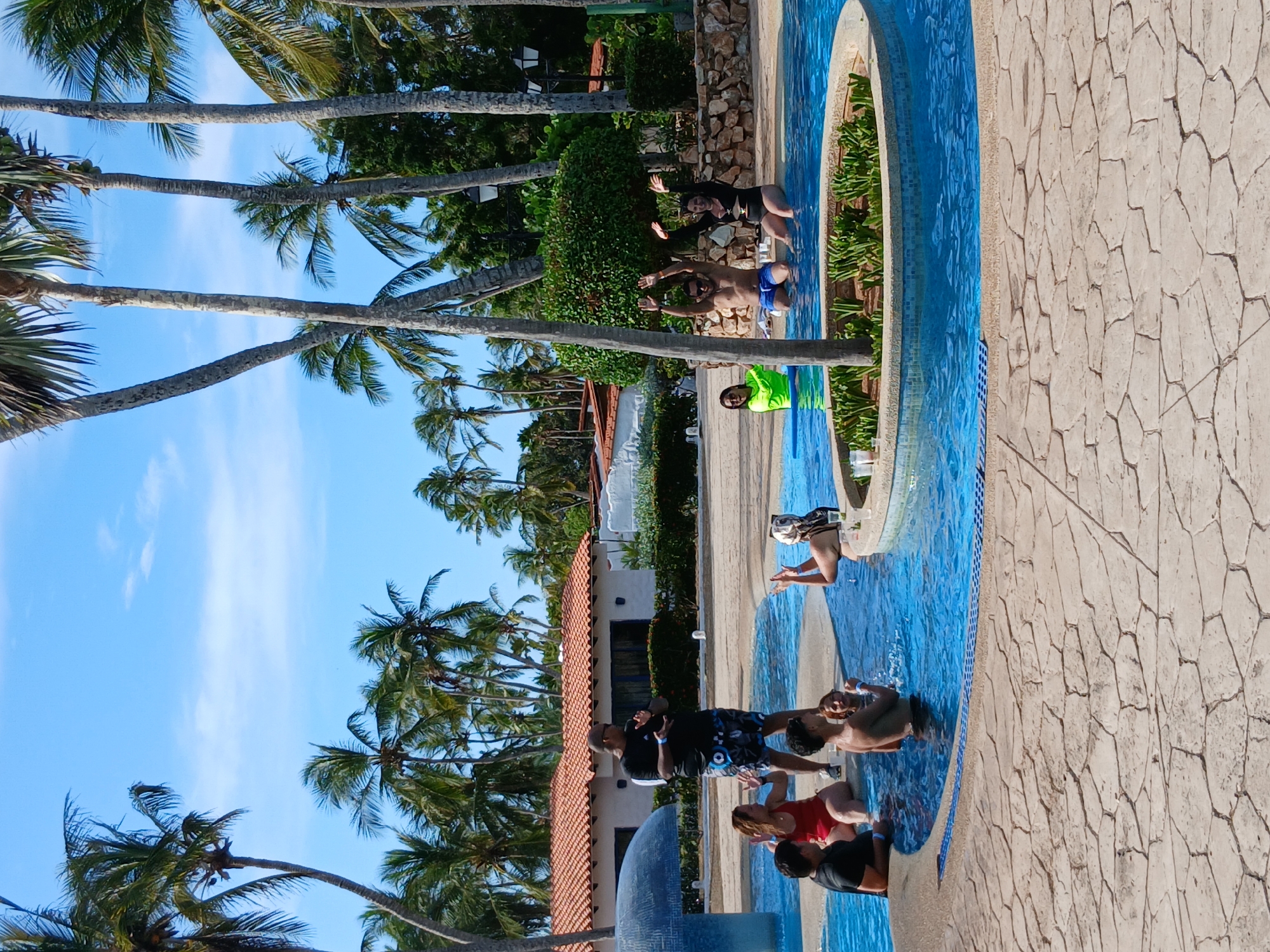 The image shows a group of people in and around a swimming pool, which appears to be part of a resort or hotel. Palm trees are visible in the background, suggesting a tropical location. The sky is blue with some scattered clouds. The people are engaged in various activities, such as swimming and relaxing. The pool is lined with blue tiles and has a stone-paved area around it. The location is Playa El Agua, Venezuela.