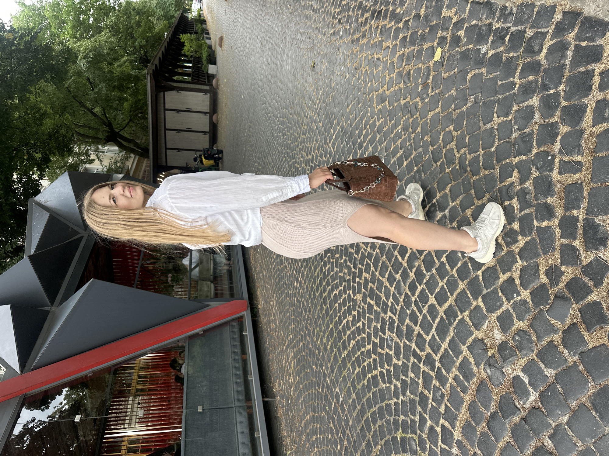 In Ternopil, Ukraine, a young blonde woman wearing a white shirt, beige skirt, and white sneakers walks on a cobblestone street. She carries a brown handbag with a chain strap. Behind her, a modern-looking building with a dark gray and red facade, and large windows, is visible. Further down the street, there are trees, a covered outdoor seating area, and a parked scooter. The overall scene appears to be an outdoor urban setting.