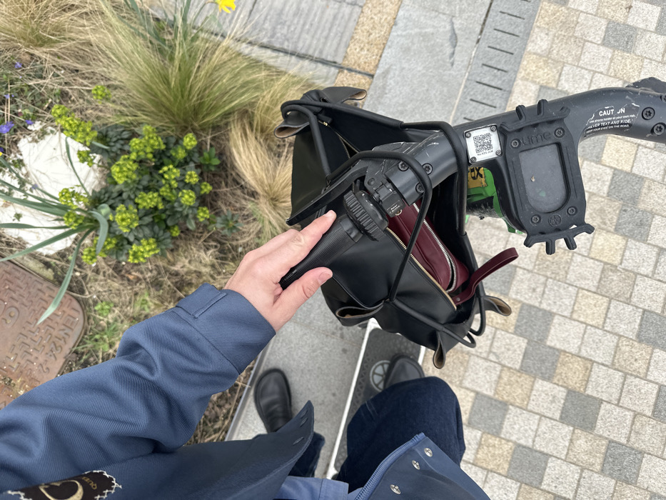 An overhead photograph depicts a partial view of an individual standing on a paved surface, interacting with items positioned on an electric scooter. The person is wearing a blue jacket/coat over a lighter blue garment and dark blue jeans. Their right hand is inserted into a black, open-top bag, which contains a maroon-colored wallet or pouch and a black electronic device, possibly a camera with a lens. The bag rests on the handlebars of a black Lime electric scooter, identified by a white "Lime" sticker, a visible QR code, and partial text reading "CAUTION". The paved ground consists of alternating light and dark grey square pavers. Adjacent to the pavement is a narrow strip of landscaping featuring a plant with bright green clustered flowers/buds, dry grasses, and a partial view of a yellow daffodil. Embedded within this landscaped area is a square metal manhole cover embossed with the letters "BT" (British Telecom), "24", and "TEL". The presence of the "Lime" scooter and the "BT" branded manhole cover confirms the location as the city of London, United Kingdom.