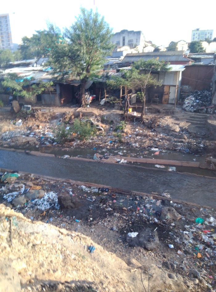 The image displays an informal settlement in Nairobi, Kenya. The foreground consists of dry, uneven earth extensively covered with diverse solid waste, including plastics, paper, and general debris. Across the mid-ground, a concrete-lined channel, approximately 3-4 meters wide, contains murky, dark-colored water with visible floating refuse. Both banks of this channel are heavily saturated with accumulated trash and organic matter.
Beyond the channel, a dense aggregation of single-story, informal structures is present, constructed primarily from corrugated galvanized iron sheets and scrap wood, with corrugated roofs and earthen foundations. Several trees with green foliage are interspersed among these dwellings. A red and white motorcycle is parked adjacent to one structure on the left side of this cluster. Towards the right, a large mound of dark, unidentifiable material, consistent with discarded tires or rubber, is piled next to another dwelling, alongside a rough set of steps descending towards the channel.
The distant background reveals urban development, including a modern multi-story building on the far left and a large, light-colored building with a distinct roofline further right, positioned above the informal settlement. The scene is illuminated by natural daylight. No human figures are discernible within the frame.