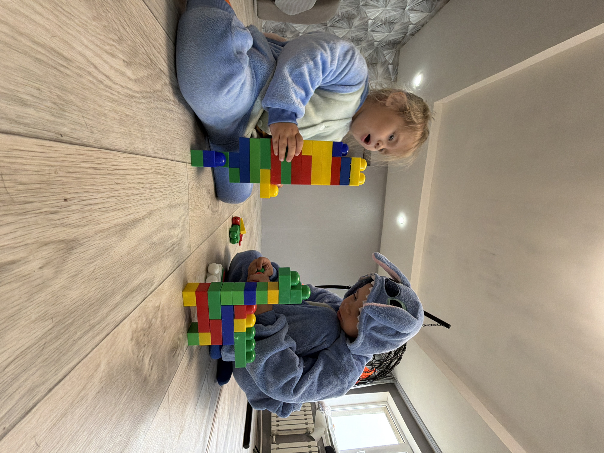 Two children are playing with plastic building blocks on a light-colored wooden floor inside a room. The first child, positioned at the top of the image, is fair-skinned with blond hair and is wearing a light blue pajama set. The second child, located at the bottom of the image, is wearing a blue Stitch character onesie. Both children are holding assembled blocks, which resemble toy guns. The room has a modern interior, featuring a geometric patterned wallpaper on the ceiling and recessed lighting. A window is visible in the background.