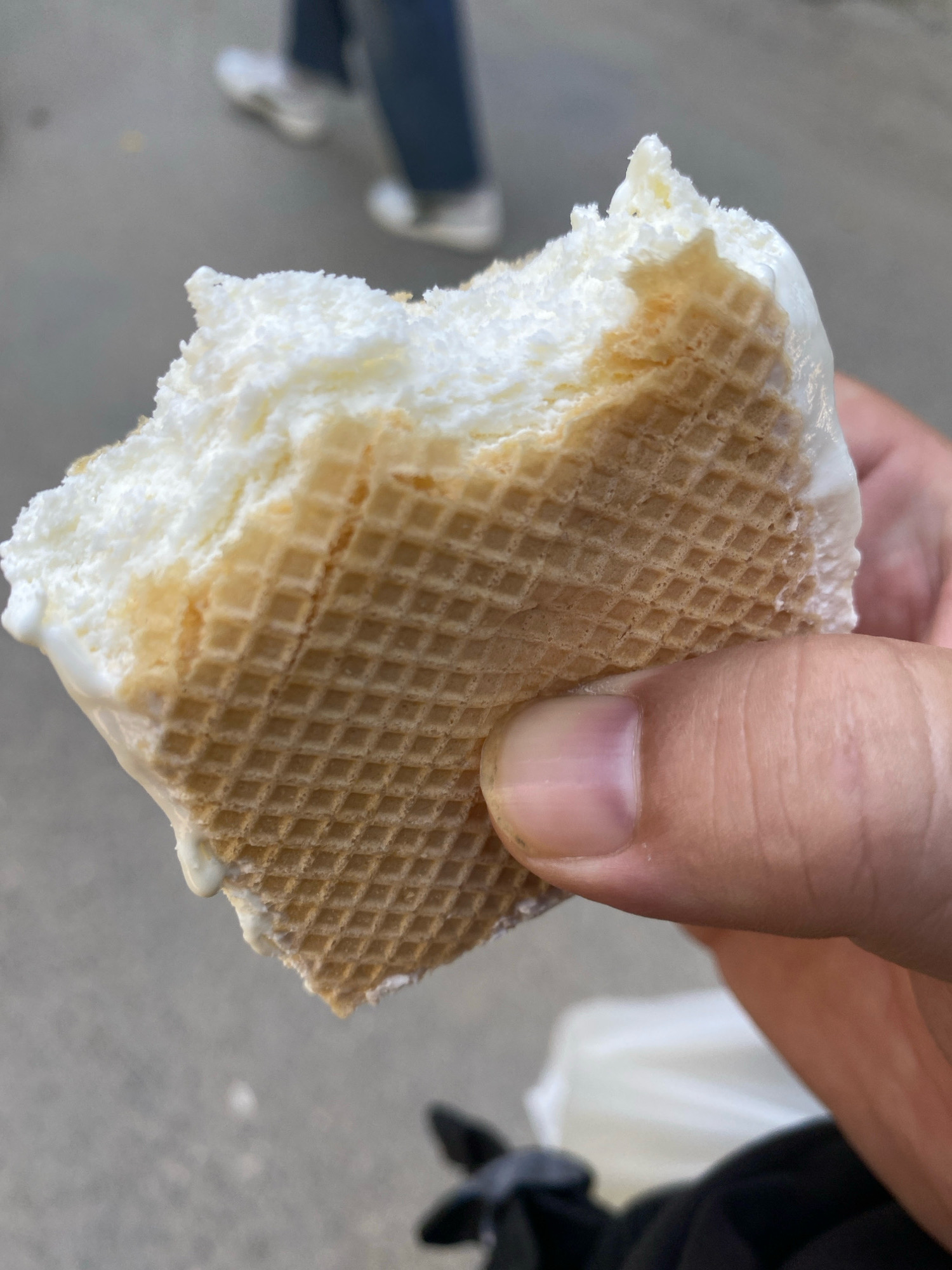 A close-up shot shows a hand holding a piece of ice cream sandwich with a bite taken out of it. The ice cream is white and appears to be melting slightly, with some caramel-colored streaks visible. The sandwich is made of two thin, square wafers with a grid pattern. The background is blurred and shows pavement and the lower legs and feet of two individuals wearing jeans and white sneakers, suggesting an outdoor setting in a city. The overall lighting indicates it is daytime.
