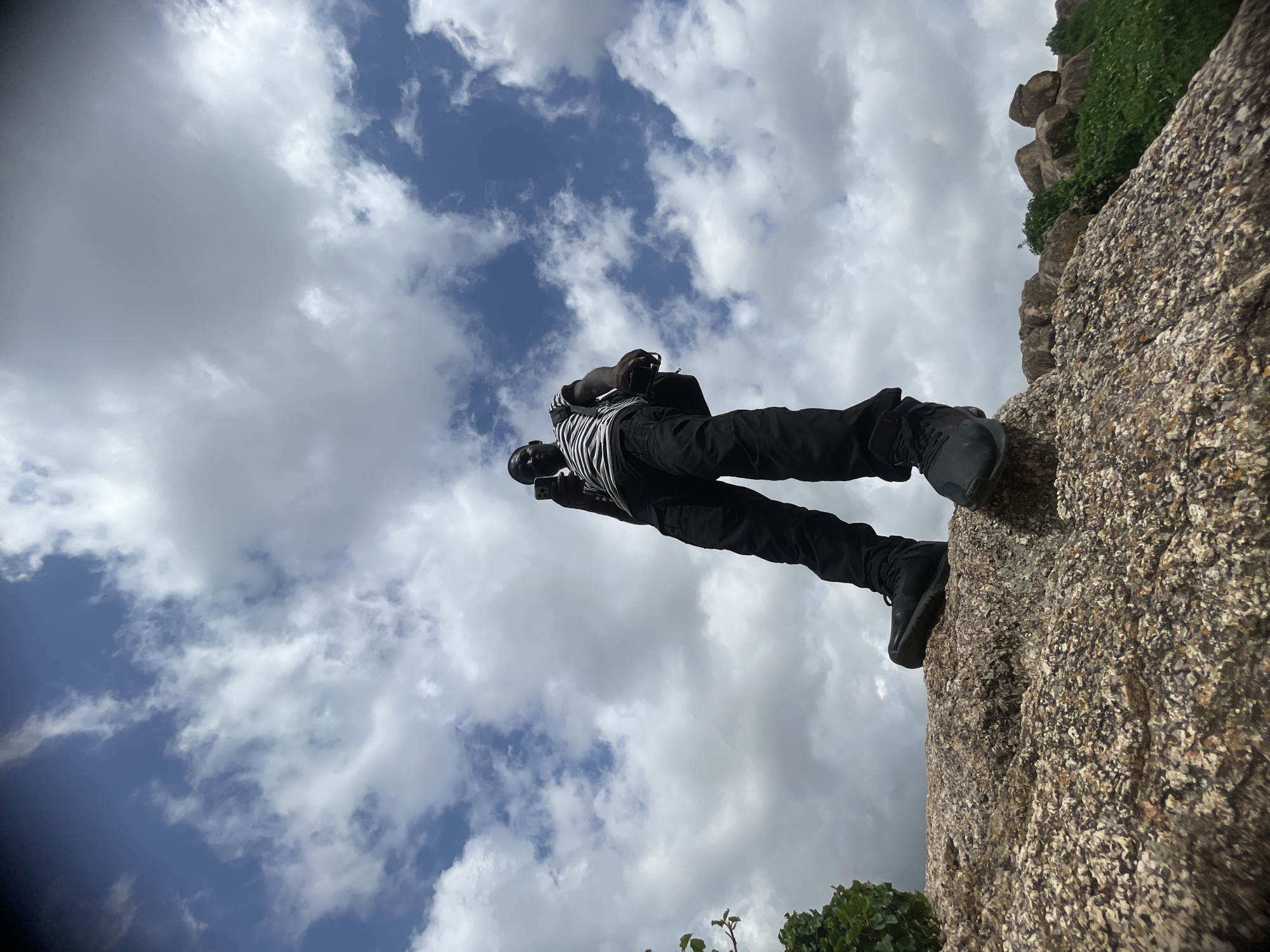 A low-angle, full shot captures a Black man standing on a rocky surface, looking upwards and holding a mobile phone in his right hand. He is dressed in a striped t-shirt, black cargo pants, and black boots. A black backpack is slung over his left shoulder. The sky above is filled with dramatic, white clouds against a blue backdrop. The foreground is dominated by the textured surface of the rock he stands on, with hints of green foliage and other rocks visible in the background. The overall scene suggests an outdoor, possibly remote, location.