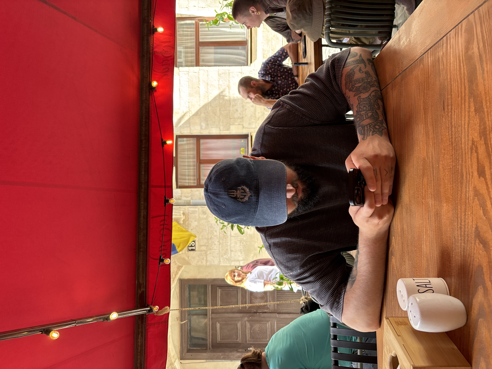 The image depicts a group of people at an outdoor cafe in Lviv, Ukraine. The cafe is covered by a red awning with string lights. A man with a beard and a baseball cap is seated at a wooden table, resting his arm with a tattoo on the table. A salt and pepper shaker are also on the table. Other people are seated at tables in the background. The background features a stone building with windows and a door.