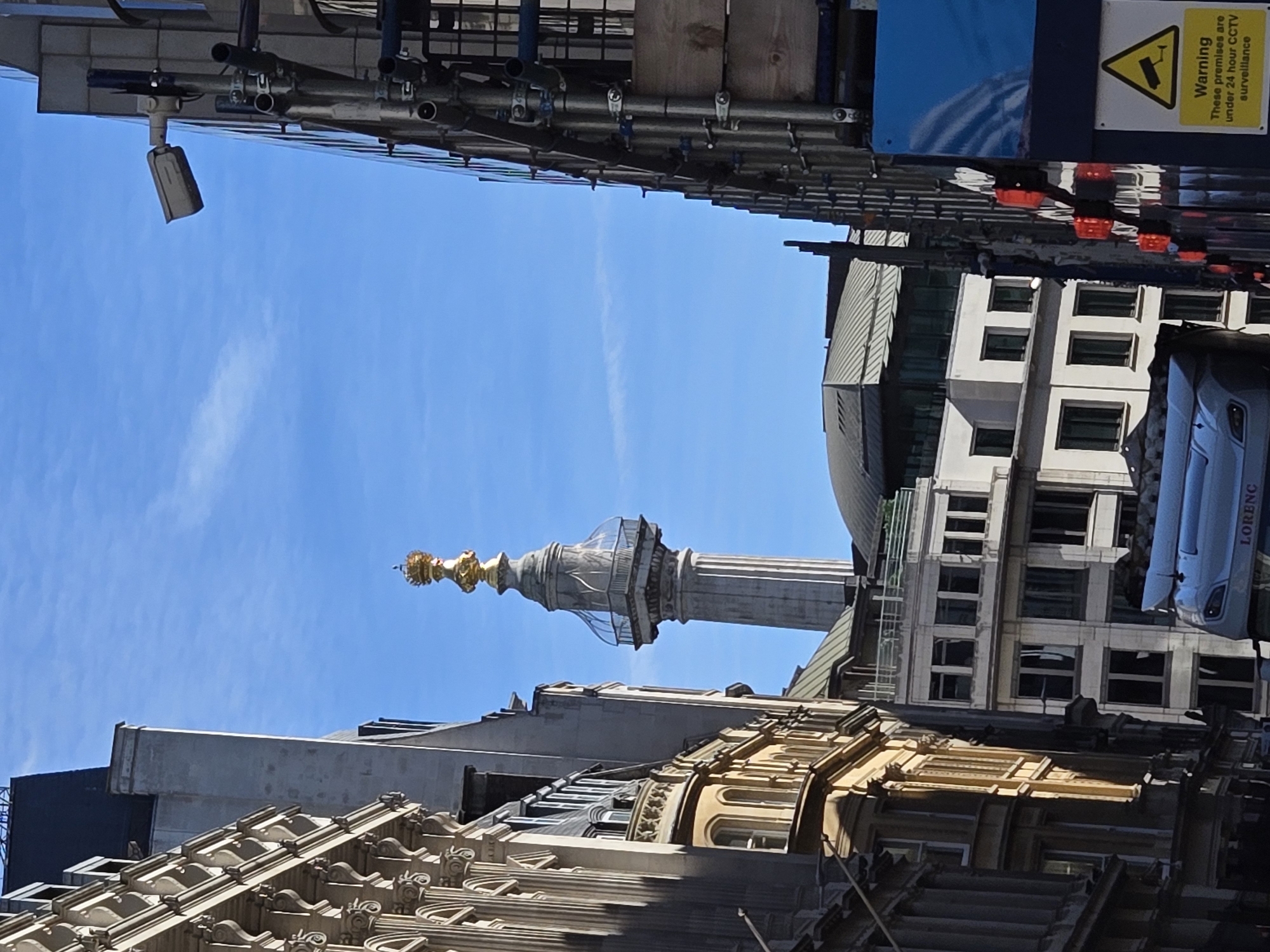 The image depicts a street scene in the City of London, United Kingdom, centrally featuring The Monument to the Great Fire of London. The Monument, a tall, fluted grey stone column topped with a gilded urn and flame, is visible against a clear blue sky, with a glass-domed viewing platform below its pinnacle.
To the left, an ornate, multi-story building with classical architectural details and numerous windows, rendered in a light stone color, partially frames the view. To the right, a building is encased in extensive metal scaffolding. A grey, cylindrical CCTV camera is mounted high on a pole attached to this scaffolding. Below, a blue construction hoarding displays a yellow warning sign that reads "Warning: These premises are under 24 hour CCTV surveillance."
In the lower center, the front section of a light-colored vehicle, possibly a van or small truck, with the name "LORENC" visible, is partially captured. Additional multi-story buildings of varying architectural styles are discernible in the background and middle ground, including one with a dark, domed roof. The scene is illuminated by daylight under a bright blue sky with minimal, wispy clouds. No individuals are clearly visible.