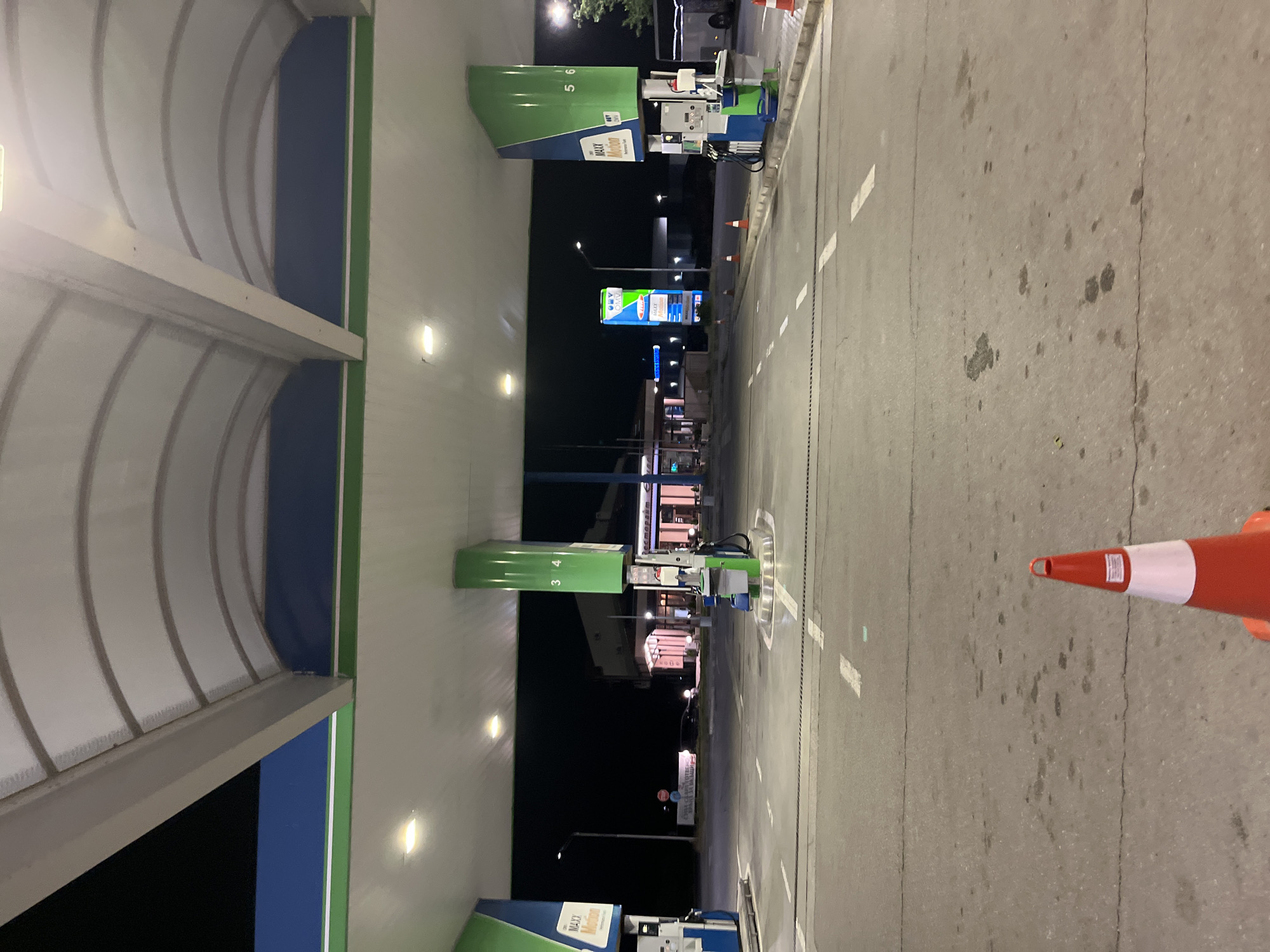 The image depicts a gas station in Popovitsa, Bulgaria at night. There are multiple gas pumps with green and blue canopies. A sign displaying fuel prices is visible in the distance. The foreground shows an asphalt road with lane markings and an orange traffic cone. The gas station is well-lit, contrasting with the surrounding darkness.