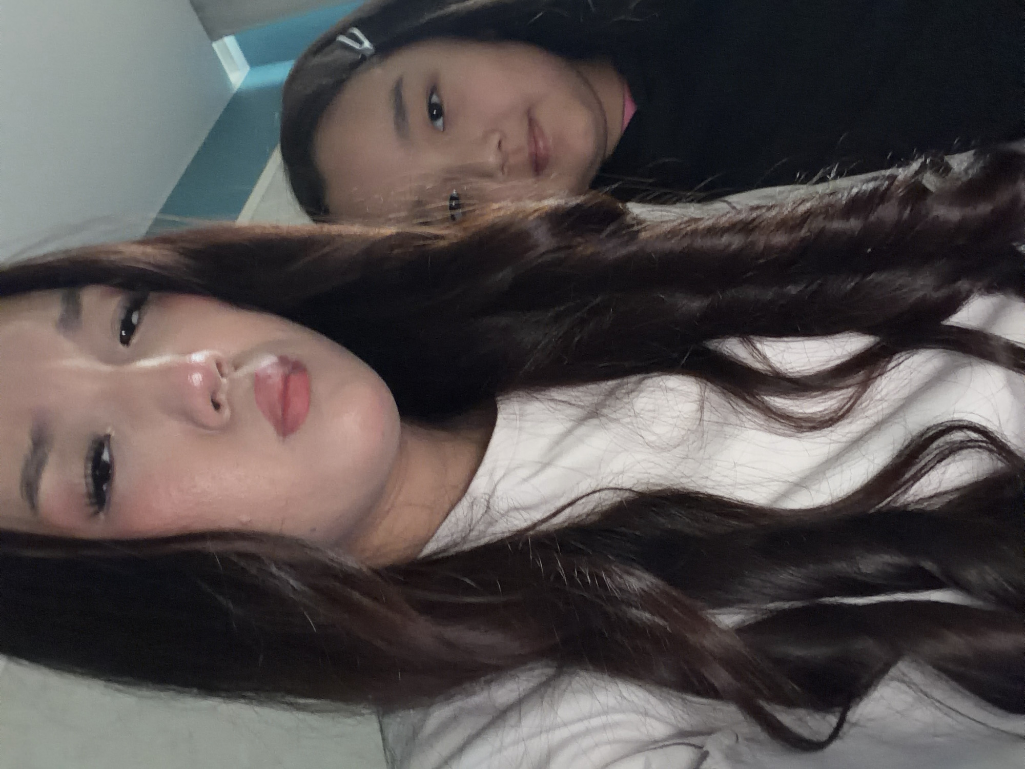 Two individuals, likely female, are captured in a close-up, selfie-style photograph. The person in the foreground, on the left, has long, dark, wavy hair and is wearing a white top. Their face is partially visible, with makeup including eye shadow and lipstick. The person in the background, on the right, has dark hair with a silver hair clip and is wearing a black top. Their face is also visible, looking towards the camera. The background appears to be indoors, with a wall and ceiling visible. The lighting suggests an indoor environment. The scene takes place in an unknown location within the city of Unknown.