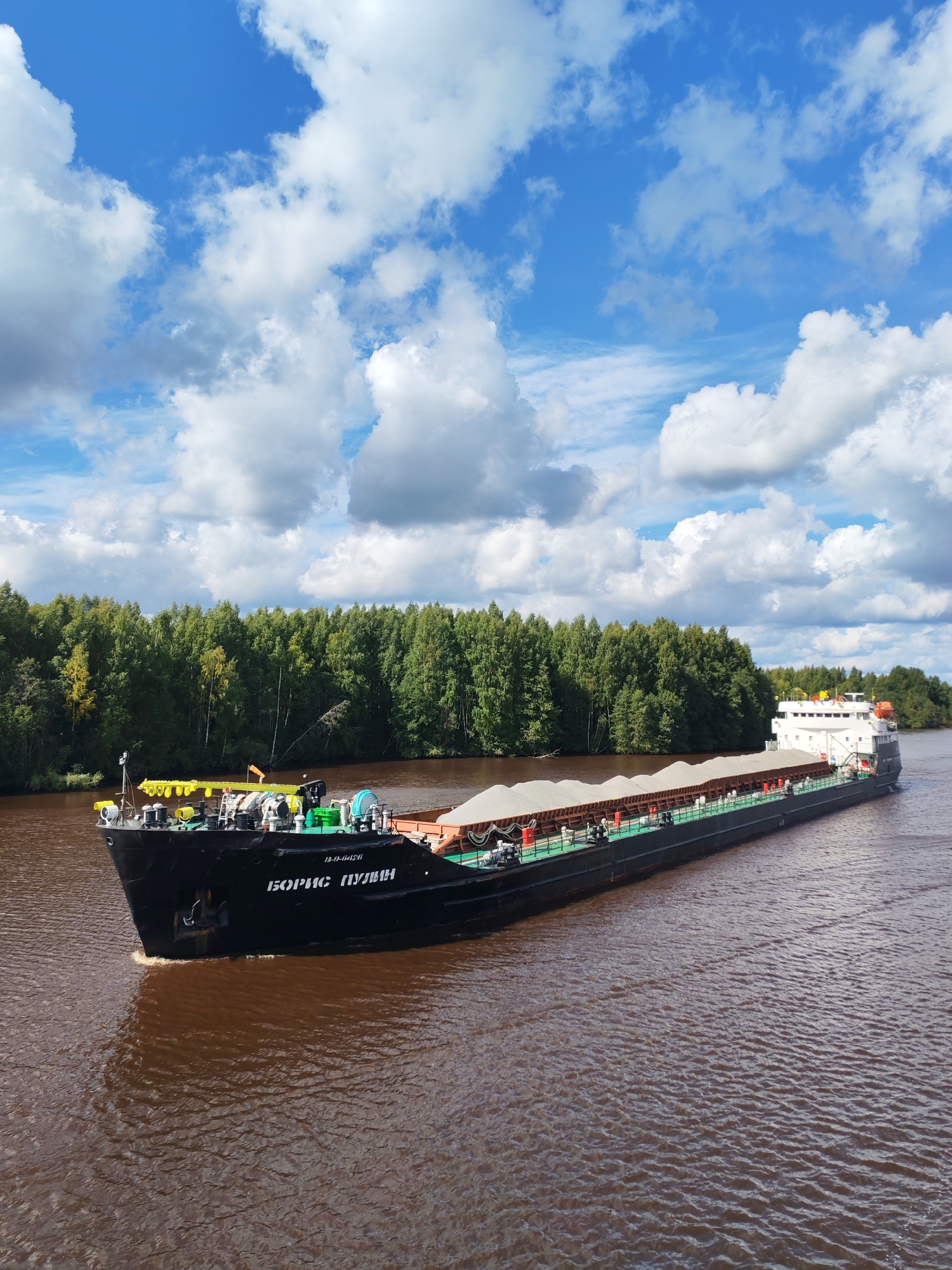 An aerial, eye-level, long shot shows a cargo barge named "БОРИС ПУШКИН" traveling down a muddy brown river. The barge is laden with white granular cargo covered by tarps, and it has a white superstructure at its stern. The riverbanks are lined with a dense forest of green deciduous trees. Above, a bright blue sky is filled with fluffy white clouds. The scene appears to be set in a rural or natural environment, possibly near Annensky Most, Russia, given the context provided.
