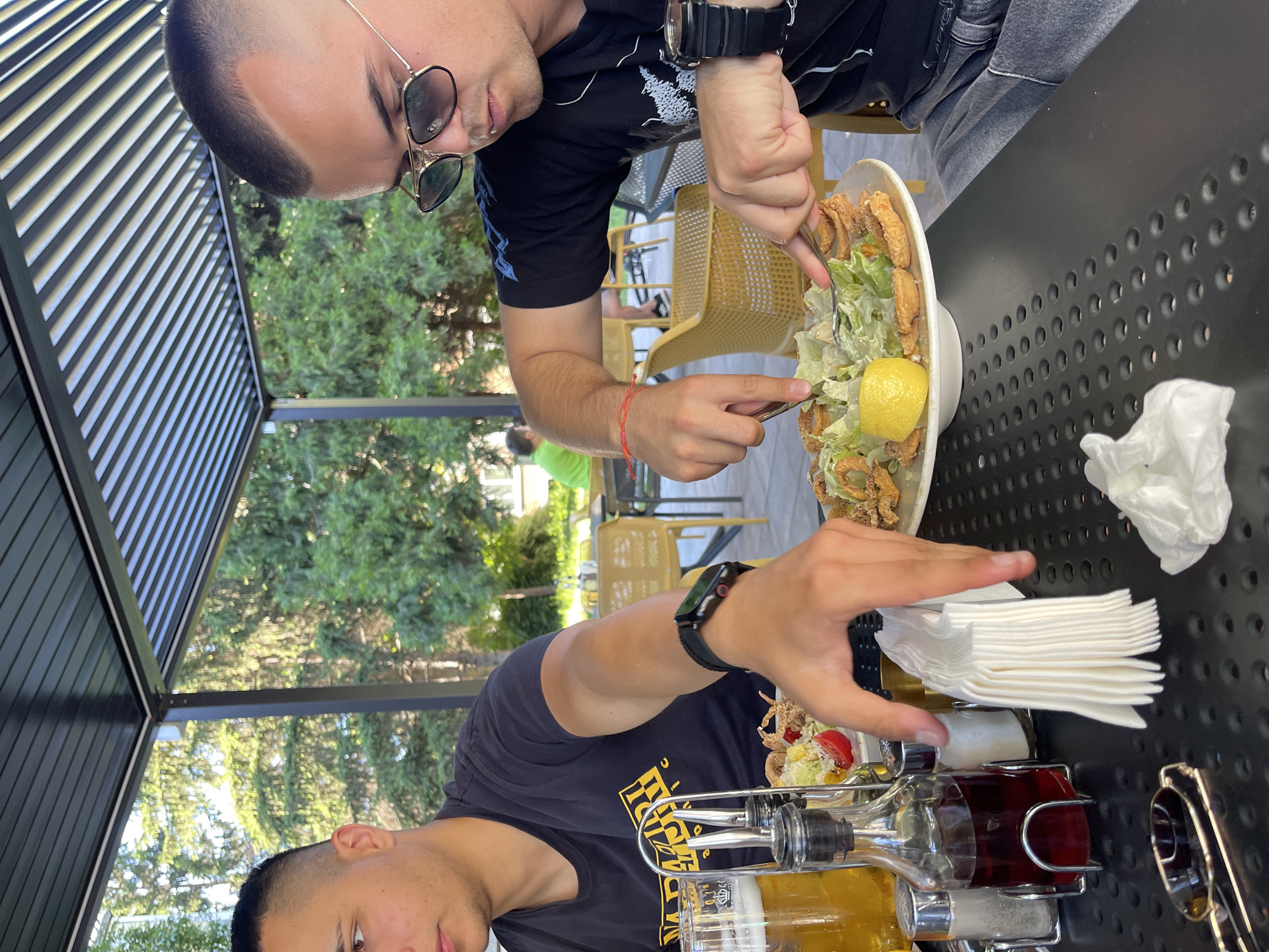 Two men are seated at a black metal table under a slatted roof structure. The man on the left is wearing a dark t-shirt with a yellow graphic design. He is looking down towards the table, with his left arm resting on the table. To his left is a glass of beer and a glass cruet with red liquid. A stack of white paper napkins is in front of him. The man on the right is wearing a dark t-shirt and sunglasses. He is actively cutting into a plate of food with a fork and knife. The plate contains a salad with breaded rings, lettuce, and a lemon wedge. A black smartwatch is visible on his left wrist. A red string bracelet is tied around his right wrist. In the background, trees and other diners are visible. The environment appears to be an outdoor dining area.