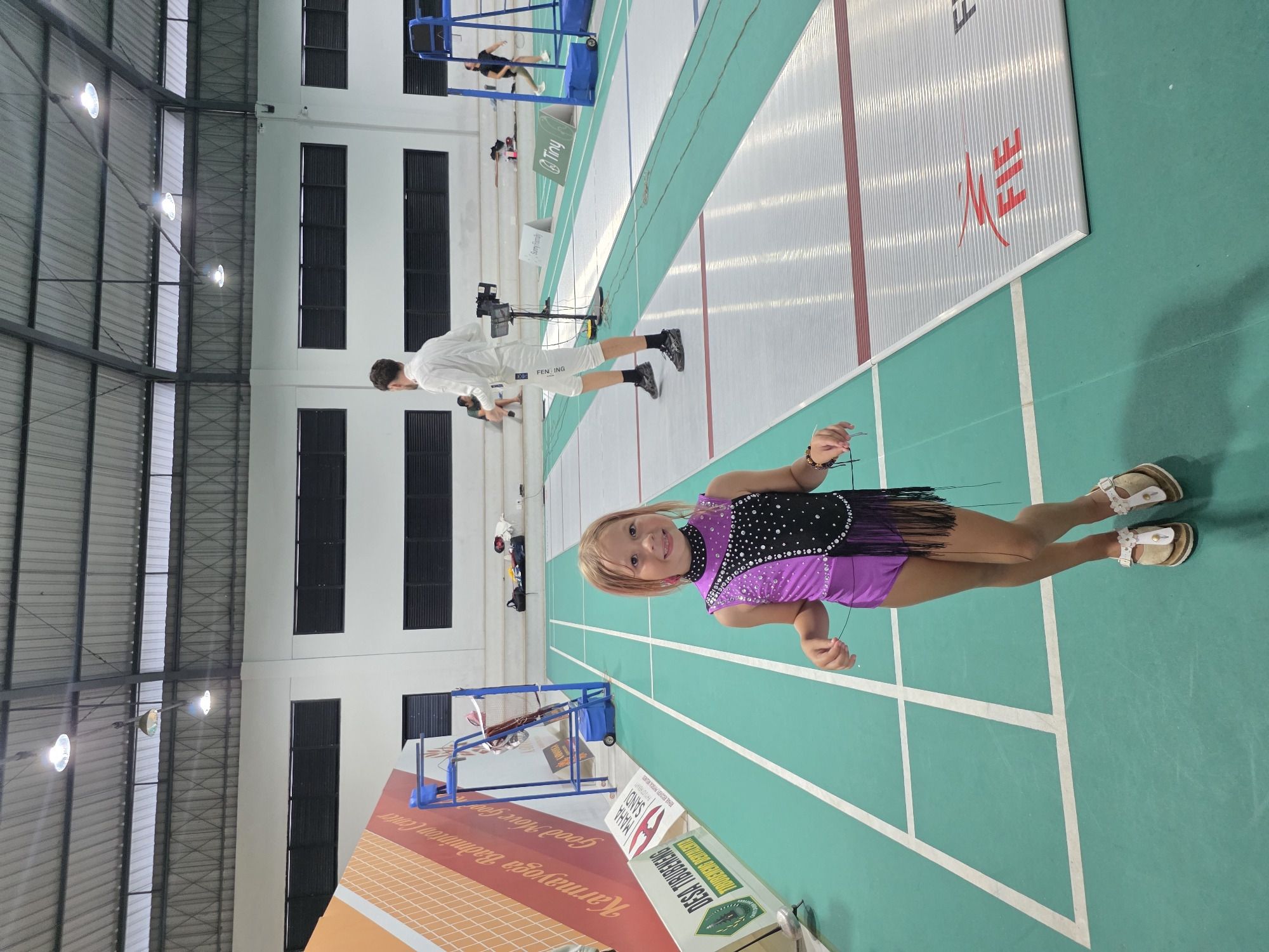 A young girl wearing a purple and black sequined dress and white sandals is standing on a green and white badminton court. In the background, a man in a white fencing suit is visible near the center of the court. Also in the background, there is a second badminton court, with what looks like a maintenance or repair lift on the sidelines. There is a large sign on the bottom left, showing the location of the Karmayoga Badminton Center in North Kuta, Indonesia. The roof of the building is high and made of corrugated metal.