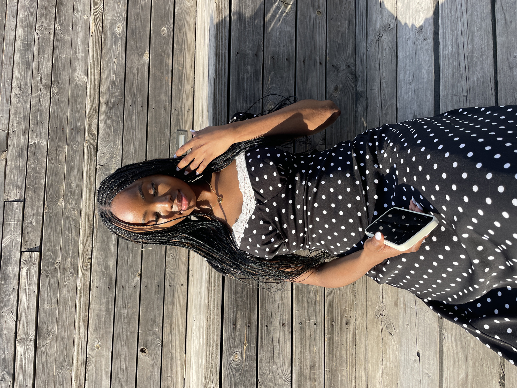 A young Black woman with braided hair is sitting outdoors against a weathered wooden wall. She is wearing a black and white polka dot dress with white lace trim. She is holding a white phone in her right hand and her left hand is near her face, touching her hair. The lighting suggests it is daytime, with shadows cast on the wall and the ground. The location is not specified as Kazan, Russia, or any other city.
