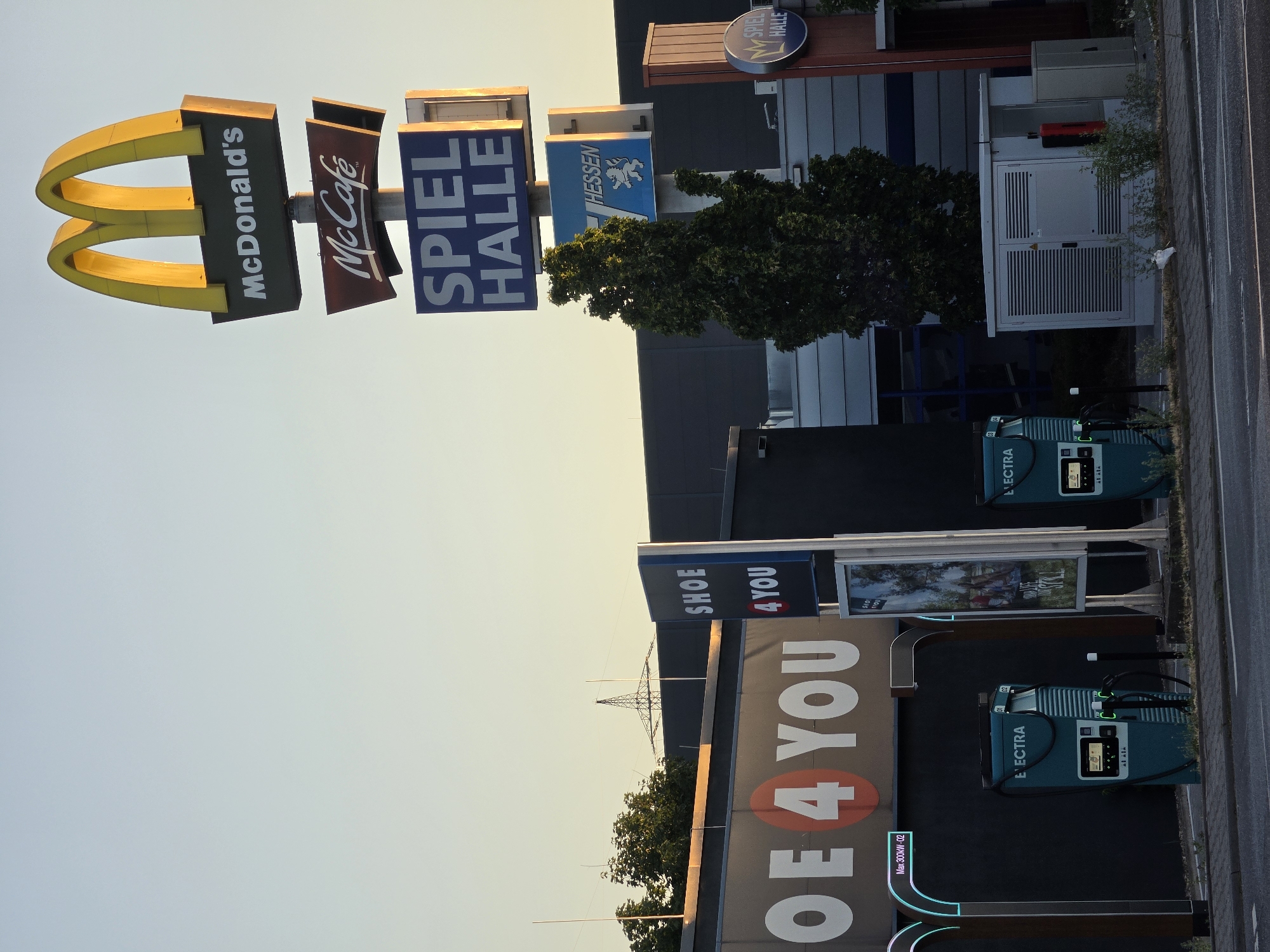 The image depicts an outdoor scene in Bischofsheim, Germany, featuring a collection of businesses and infrastructure. Prominently displayed is a tall McDonald's sign with the iconic golden arches and branding for McDonald's and McCafé. Below these, signs for "SPIEL HALLE" (Game Hall) and "HESSEN" with a lion emblem are visible. To the left, a large sign reads "OE 4 YOU," suggesting a retail store. In the foreground, there are several Electra charging stations, indicating an area with electric vehicle charging facilities. The background includes a large building, possibly a commercial complex, with a dark facade and metallic accents, and a glimpse of a power transmission tower in the distance. The environment appears to be an urban or commercial area, possibly an industrial park or a shopping district, with a road in the immediate foreground. The sky is clear and shows the soft light of late afternoon or early morning. There are no people visible in the image.