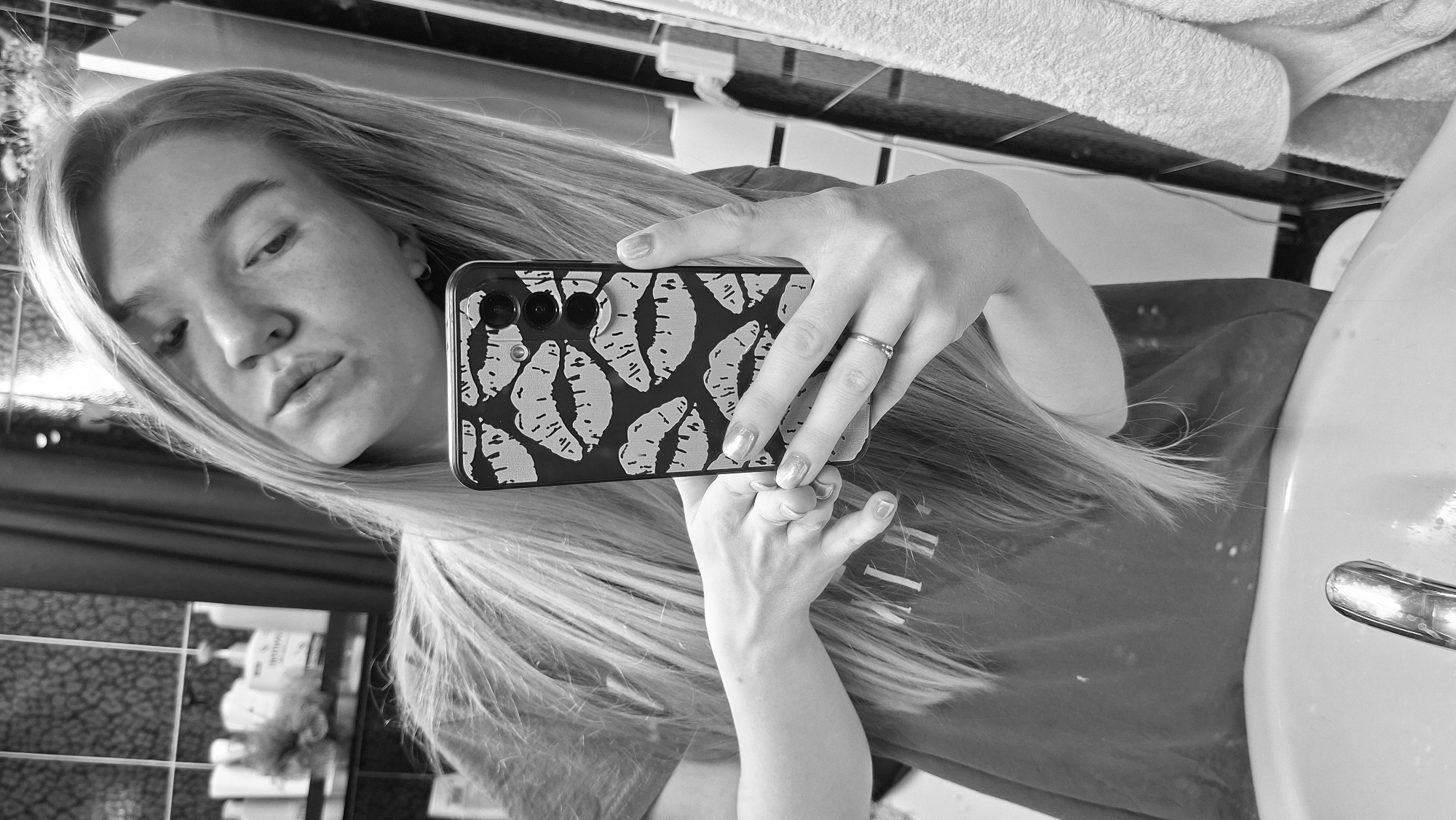 A woman with long hair is taking a selfie in a bathroom, likely indoors. The phone case is black with a lipstick kiss pattern. A sink is visible to the right, and a towel rack with towels is in the background. The exact location in the city of Chaikovsky, Russia cannot be confirmed from the image.