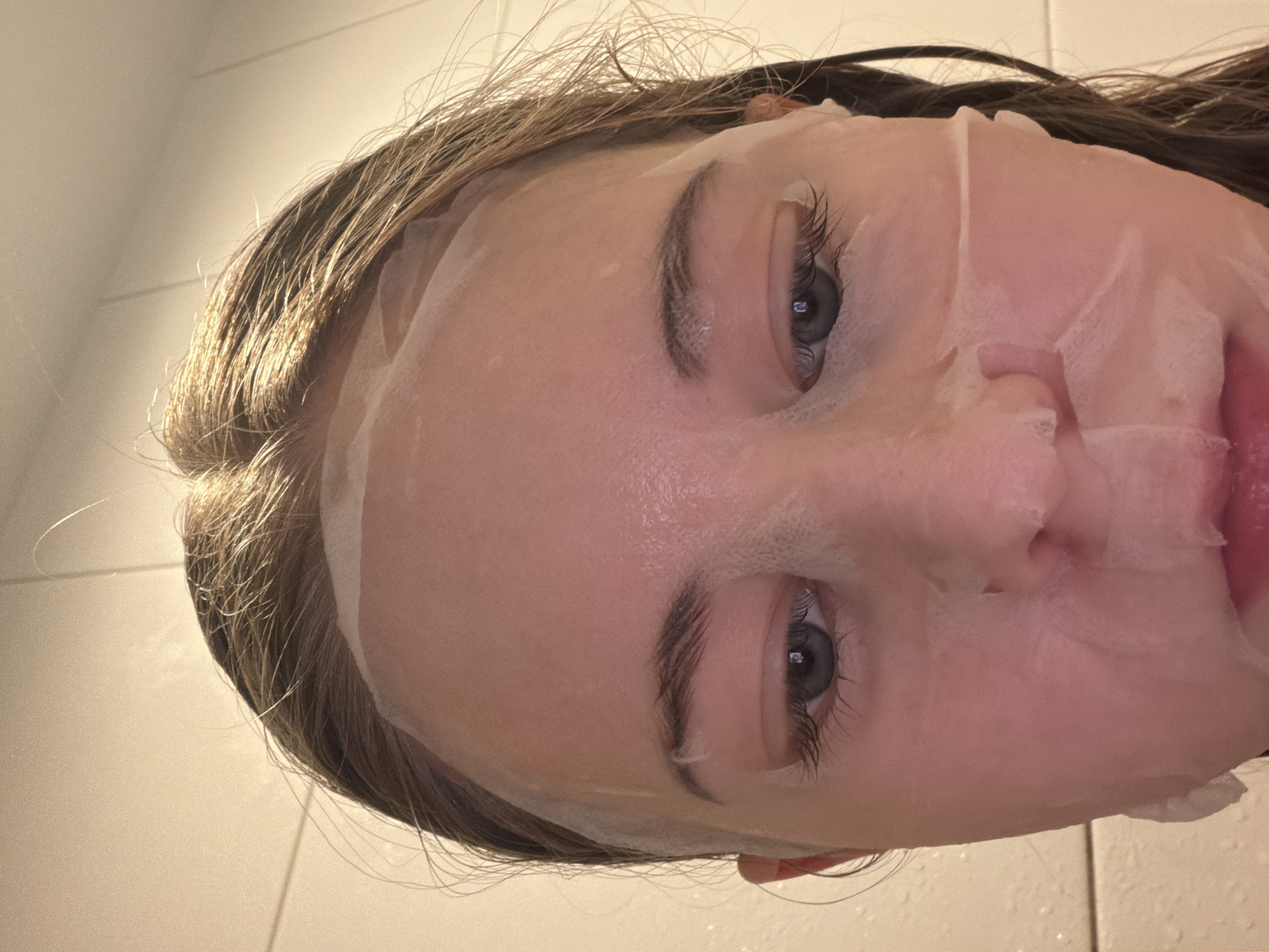 A close-up, angled shot captures a person's face adorned with a white, translucent sheet mask. The individual, appearing to be a young female, is positioned almost horizontally within the frame. Her light brown hair, with hints of blonde, cascades around her face; some fine strands are visible, catching the light on the left side of the image.
The sheet mask covers most of her facial features, with precisely cut openings for her eyes, nose, and mouth. Her eyes are open, revealing hazel or blue irises, and her long, dark eyelashes are prominent. Her eyebrows are visible above the mask. A glimpse of her pink lips can be seen through the mouth cutout on the right. The mask appears moist and adheres well to the contours of her face, indicating an ongoing skincare regimen.
The background consists of light-toned, possibly tiled, surfaces, suggesting an indoor setting, likely a bathroom. The lighting is soft and ambient, indicative of an artificial light source rather than natural daylight. The person's expression is neutral, showing no overt emotions. There is no visible text in the image. The location context for this scene is Perm, Russia.