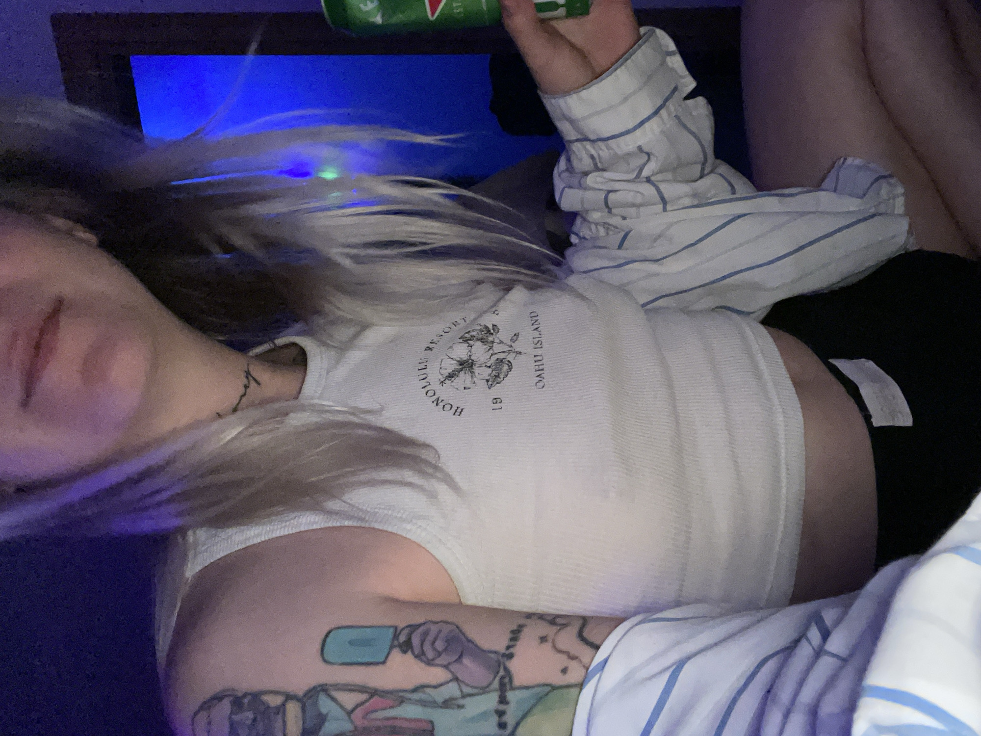 A person with light blonde hair, wearing a white tank top with "HONOLULU RESORT OAHU ISLAND 19" printed on it and black shorts, is reclining on what appears to be a bed with blue and white striped sheets. The person has a tattoo on their upper arm. They are holding a green can. A blue light source is visible in the background. The location is not discernible in this image and cannot be determined to be in Belgrade, Serbia.