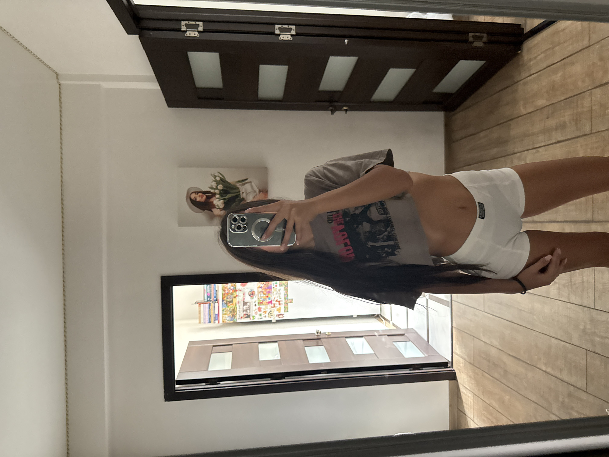 The image shows a young woman standing in front of a mirror, taking a selfie. She is wearing a gray crop top and white boxer briefs. Her long, dark hair is down, and she is holding a smartphone in her right hand. The background suggests an indoor setting, possibly a home. To her left, a door is open, revealing a hallway with more doors and some decorative items on a shelf. To her right, another door is visible. A framed picture of a woman wearing a hat and holding flowers hangs on the wall. The floor is made of light-colored wooden planks. The overall lighting is bright, indicating natural light coming from a window or being the primary light source. The location of Chișinău, Moldova is not discernible from the visual elements in the image.
