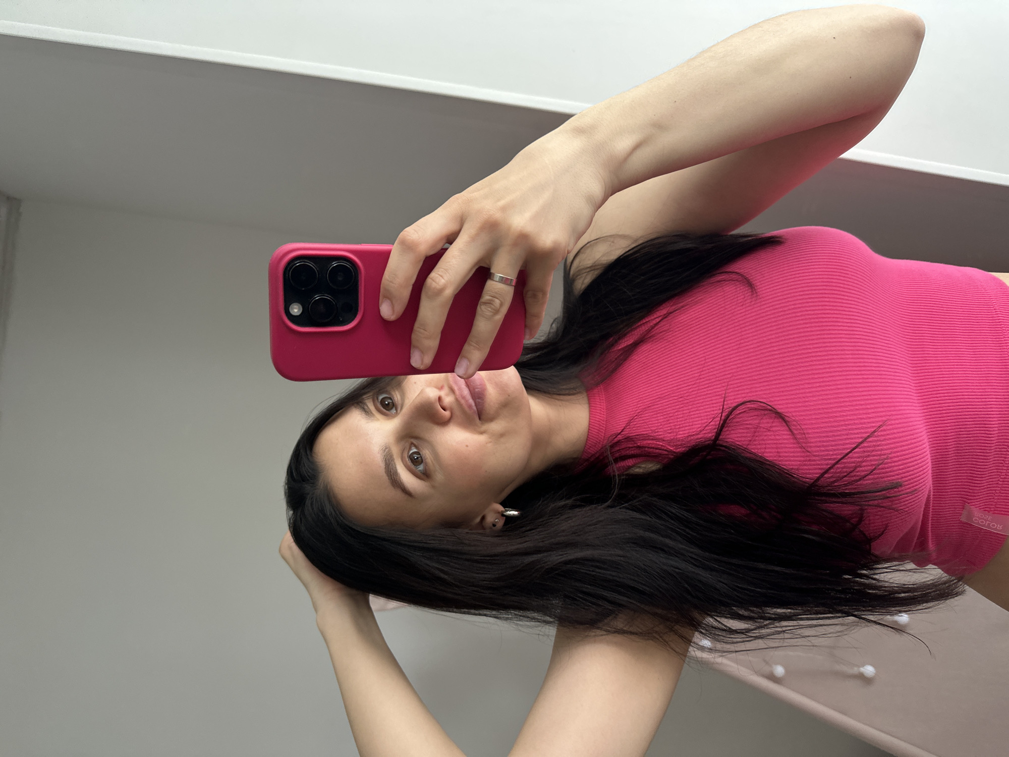 This mirror selfie, captured in Chaikovsky, Russia, features an adult woman with long, dark hair and brown eyes. She is wearing a vibrant hot pink, ribbed top, on which the text "ROSE GOTOK" is visible near the lower right edge. The woman is holding a matching hot pink smartphone, equipped with a multi-lens camera module, in her right hand, pointed towards the mirror to take the photo. A simple, flat silver-colored band ring is worn on her left ring finger. Her expression is neutral to slightly playful, with her lips softly pursed, and her gaze is directed towards the camera's reflection. Her left arm is raised, with her hand touching the back of her head.
The setting is an indoor space characterized by light-colored, possibly off-white or light grey, walls. A white horizontal surface or trim runs across the upper background. In the lower right of the image, behind the woman, small, white, spherical decorative elements are visible on a darker, possibly beige or light brown, surface, suggesting a piece of furniture or a decorative object. The lighting is bright and even, indicating an indoor daytime environment. The image itself is presented on its side, showing the woman horizontally.